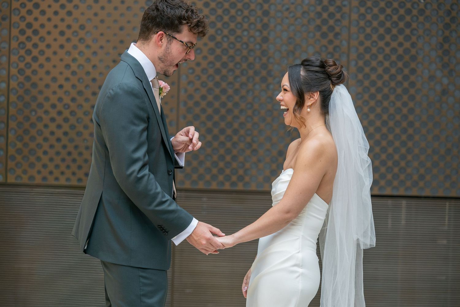 Bride in white gown and veil holds hands with groom in gray suit during romantic first look wedding moment.