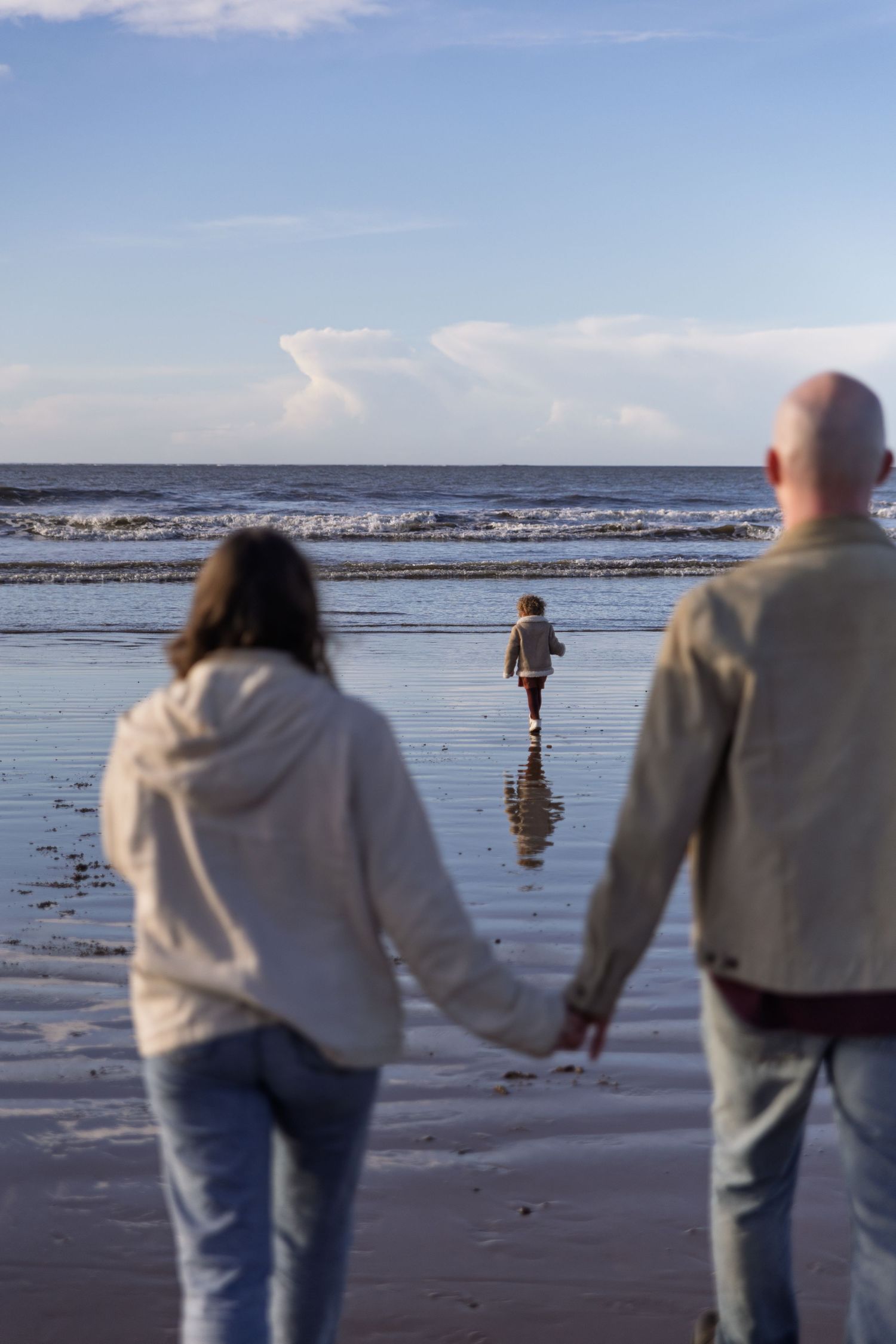 A couple holds hands while watching a child play on a reflective beach at sunset with waves and clouds in the background.