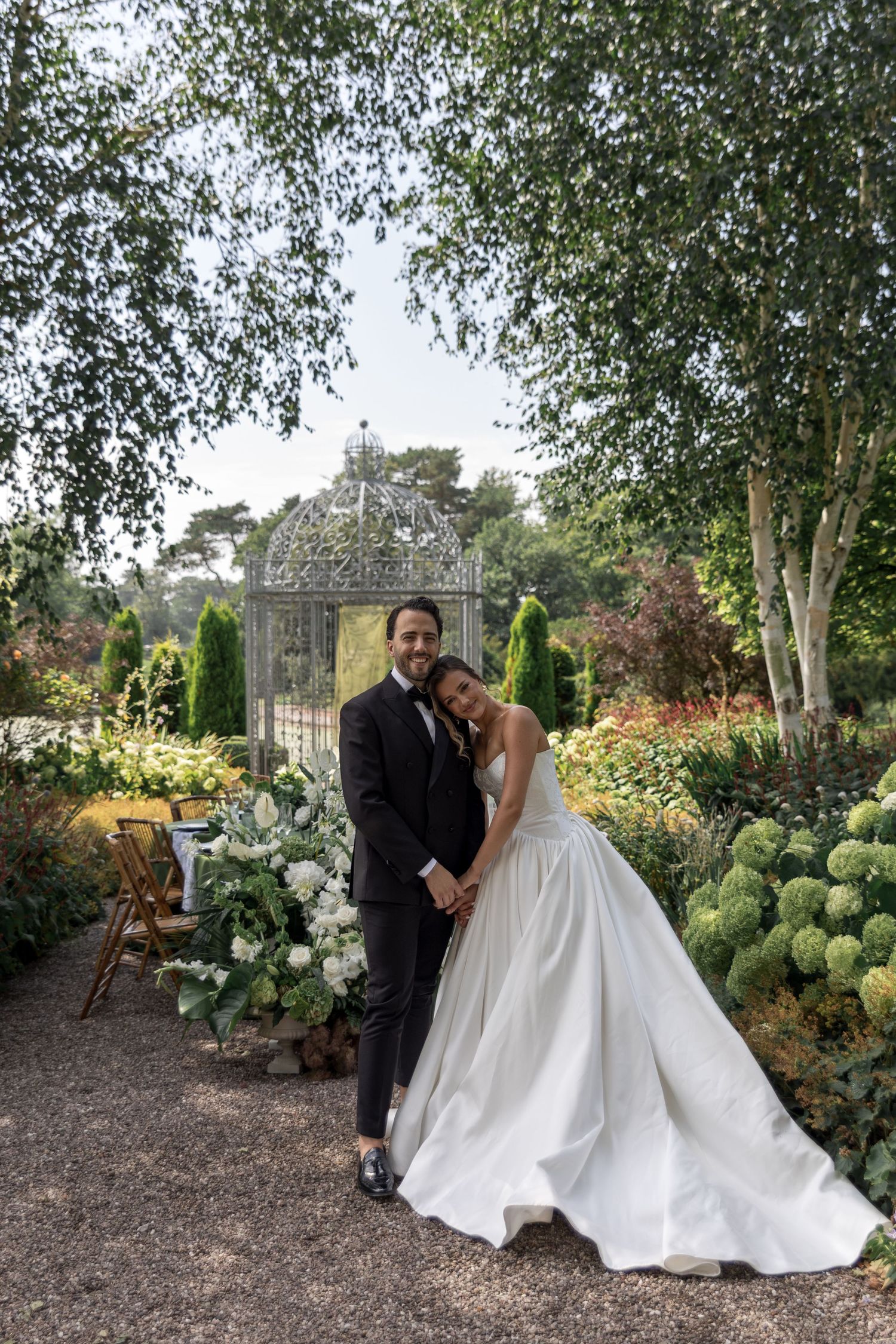 A couple in formal wedding attire poses together in a lush garden setting with hydrangeas and greenery.