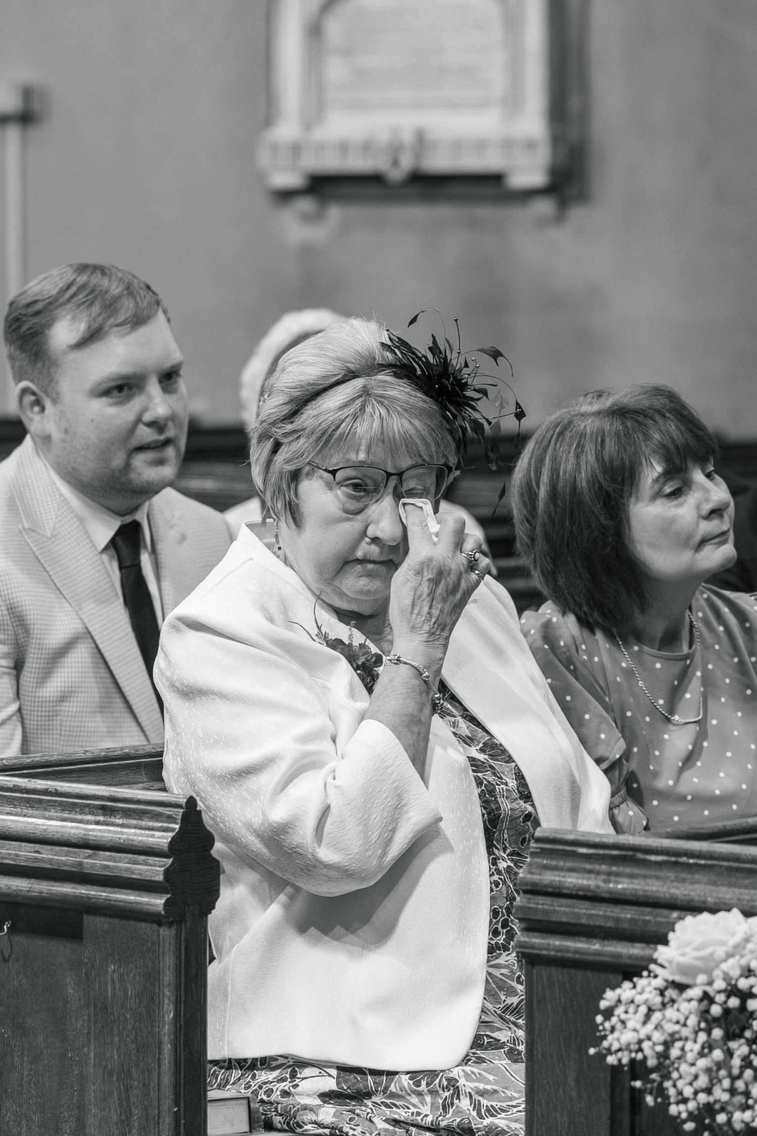 Emotional wedding guest wiping tears in church pew, black and white photo.