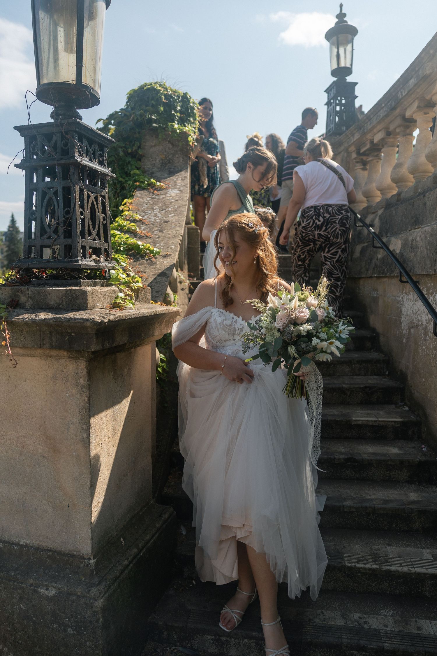 Bride in white tulle gown holding bouquet descends stone steps at elegant outdoor wedding venue, Parade Gardens, Bath