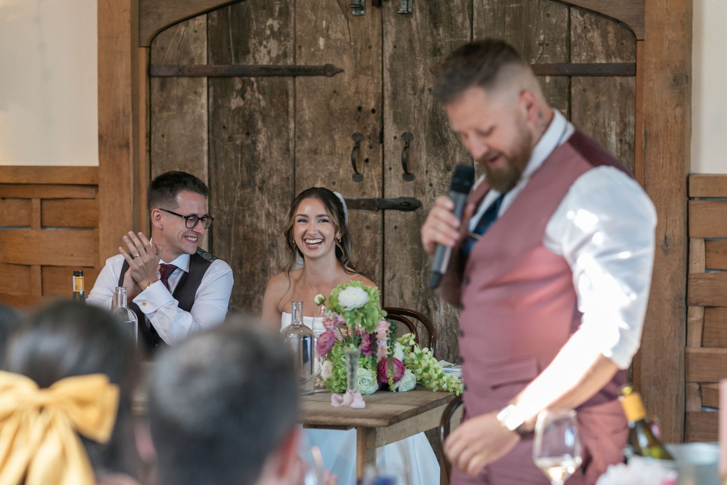 Bride and groom laugh during wedding speech by groomsman in burgundy vest at rustic barn reception.