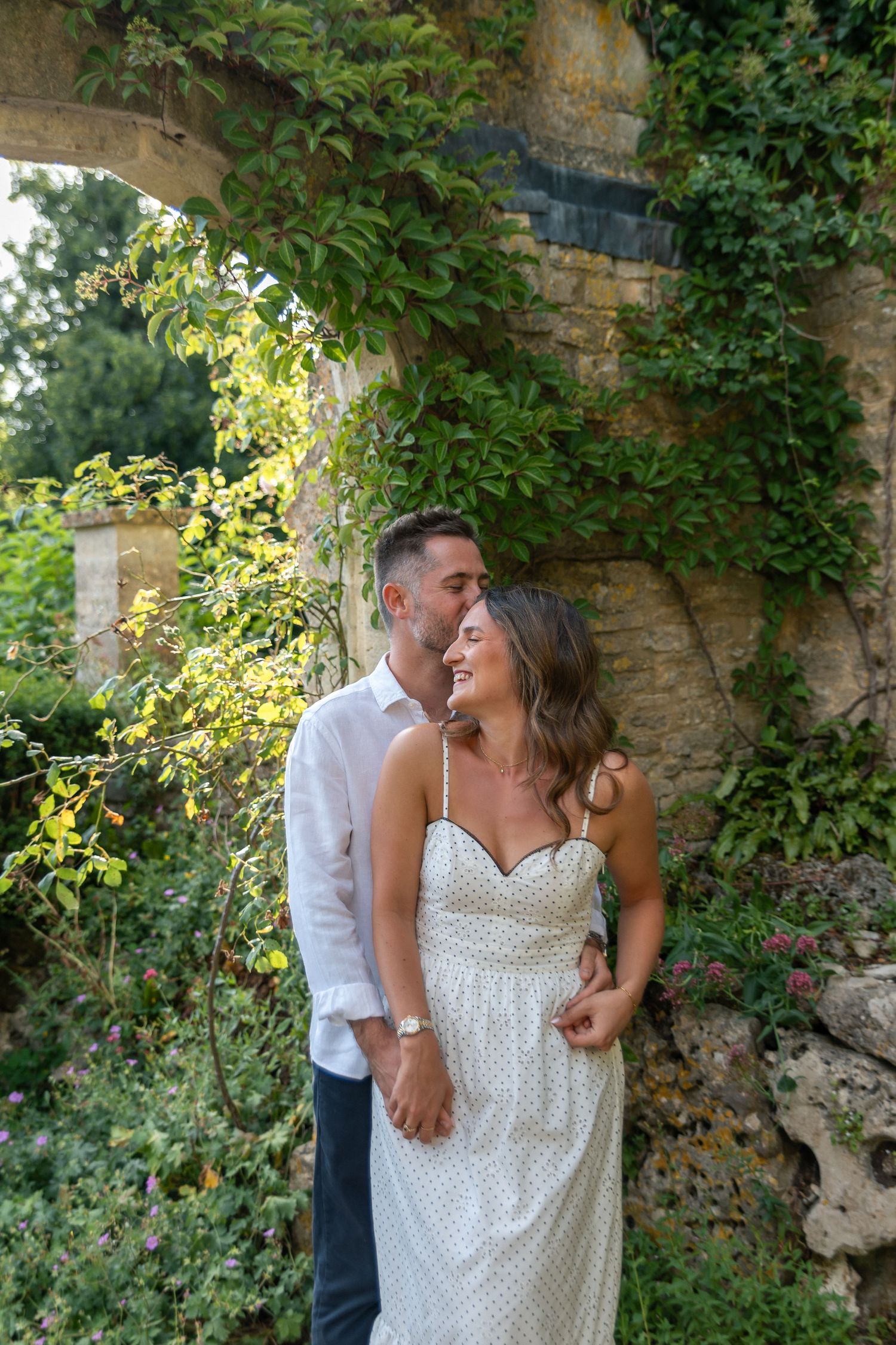 A couple shares a romantic moment under a vine-covered stone arch, the man kissing the woman's temple as she smiles.