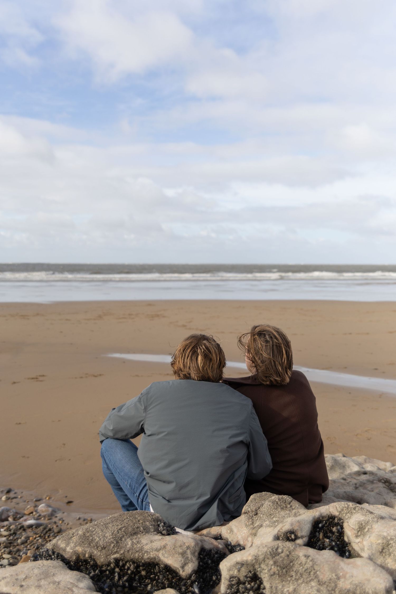 A couple rests together on large rocks by the ocean on a cloudy day.