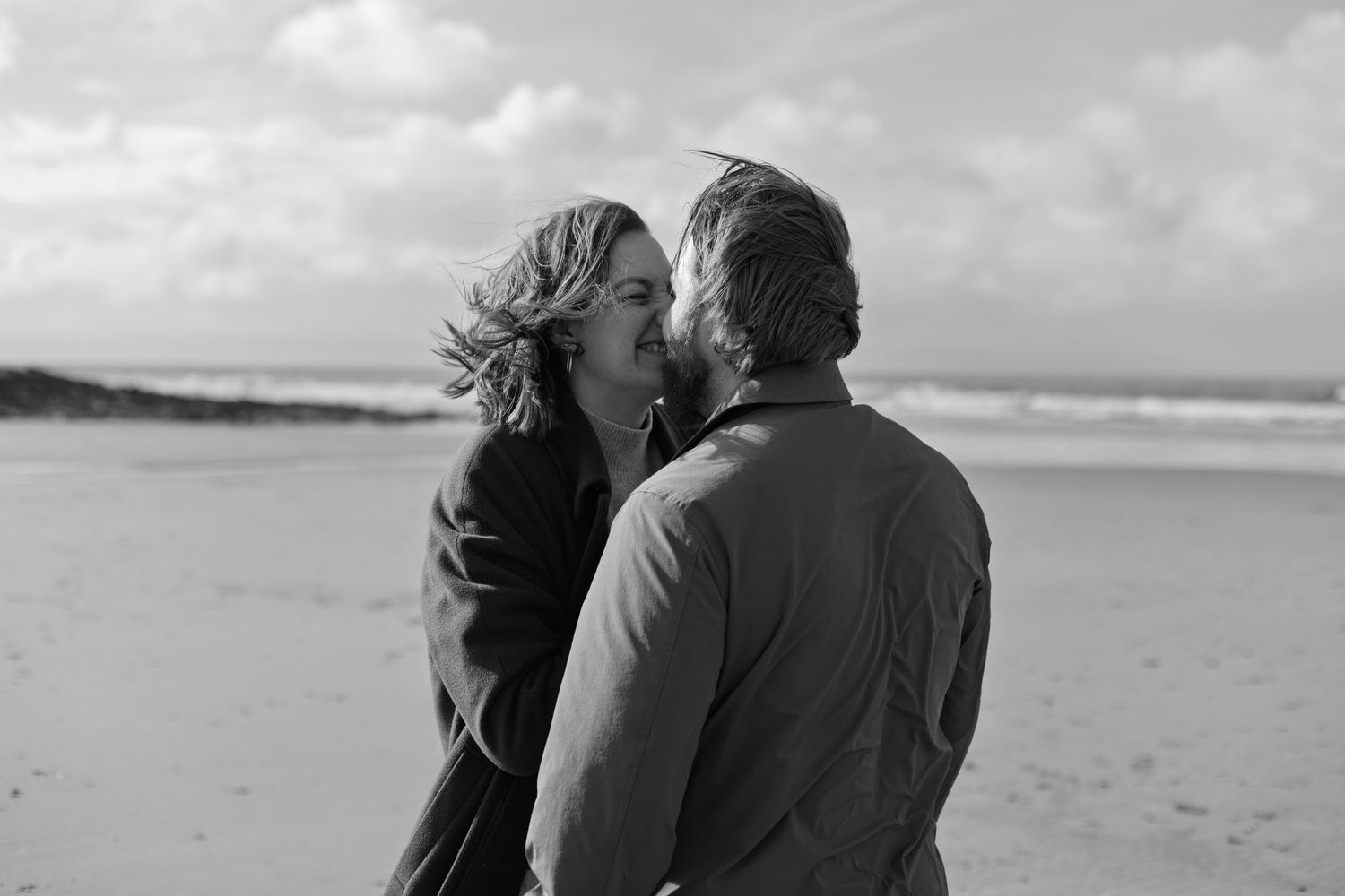 A couple shares an intimate moment while walking along a misty beach.