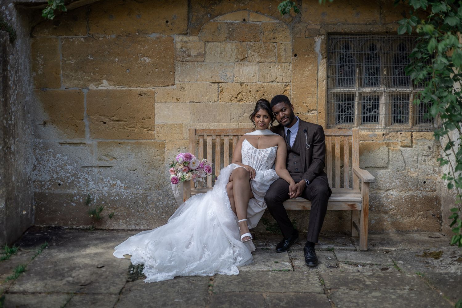 Bride and groom sitting on a wooden bench against a stone wall, bride in white gown holding pink bouquet at Sudeley Castle