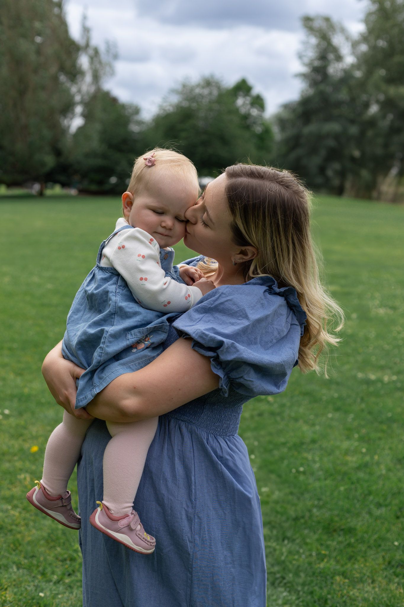 A tender family moment captured outdoors with coordinating blue outfits in a grassy park setting.