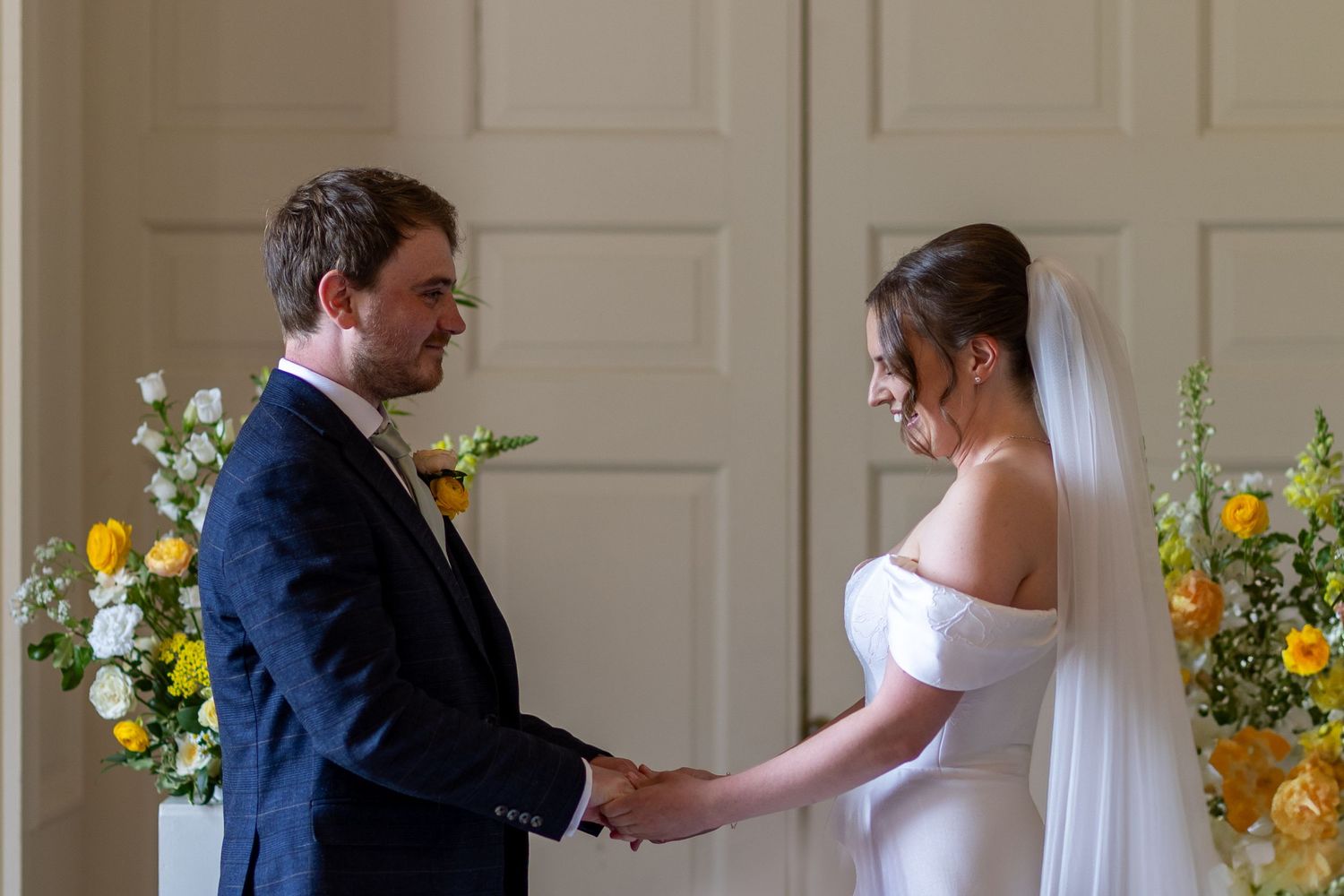 A couple exchanges vows during their wedding ceremony in front of yellow and white floral arrangements.