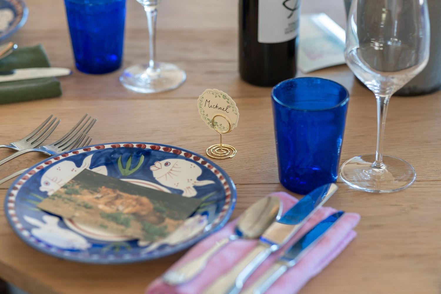 Elegant table setting with blue glassware, decorative plate, silverware, and wine bottle on wooden table.