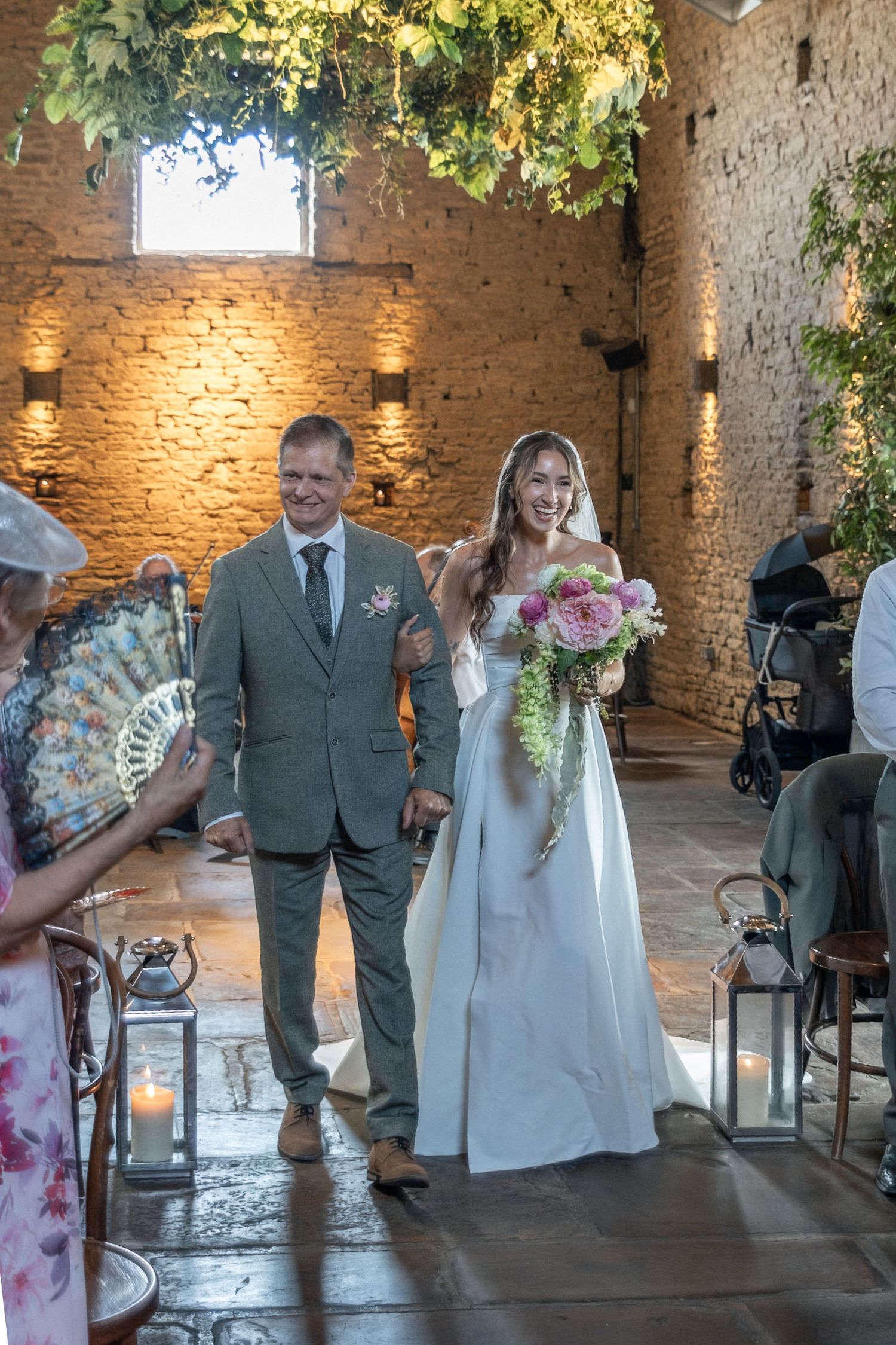 Bride in white gown holding pink bouquet walks down aisle with escort in rustic stone venue.