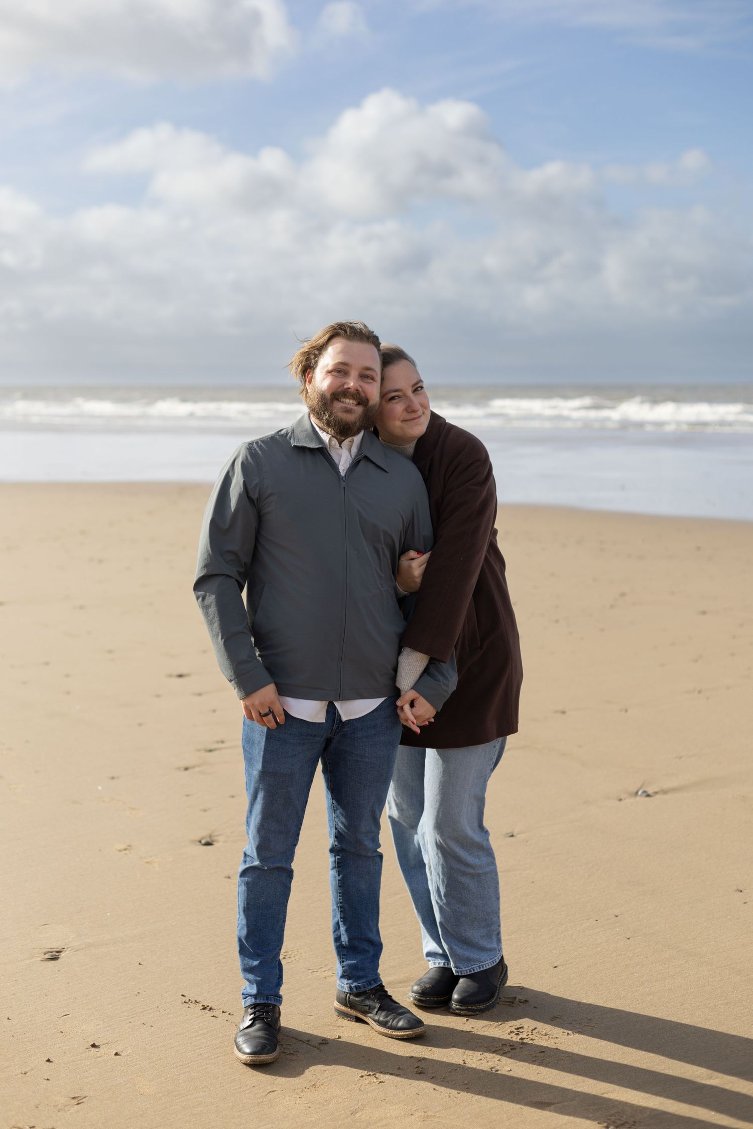 A couple stands close together laughing on a sandy beach with blue skies overhead.