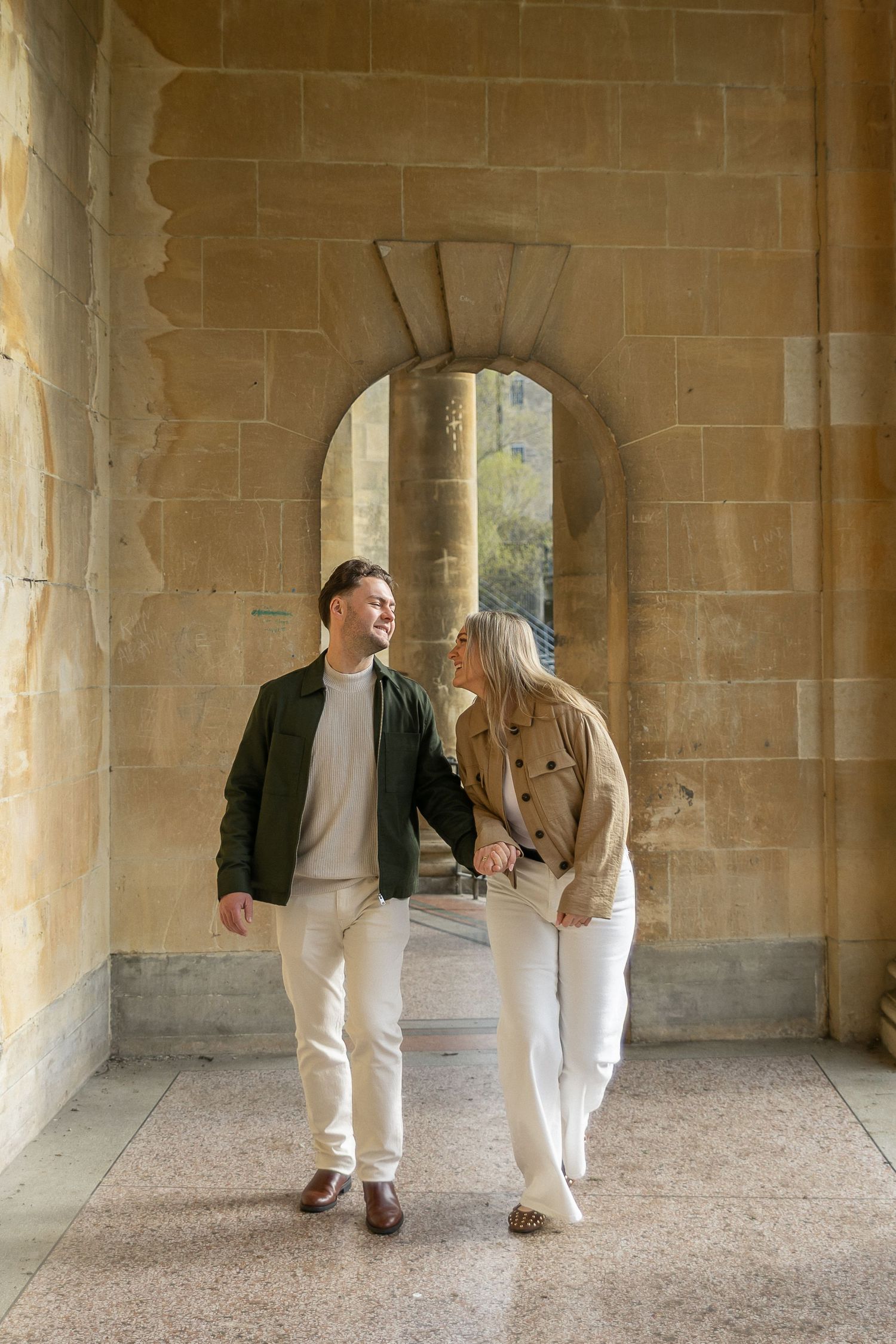 Couple holding hands and smiling at each other while walking through a stone archway corridor.
