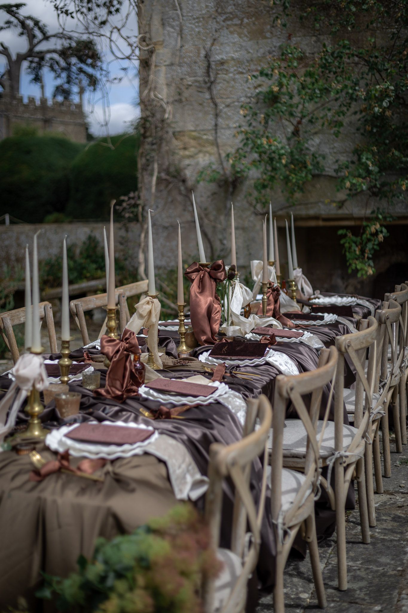 Elegant outdoor wedding reception table set with brown linens, gold candlesticks, and tall taper candles near a stone wall at Sudeley Castle