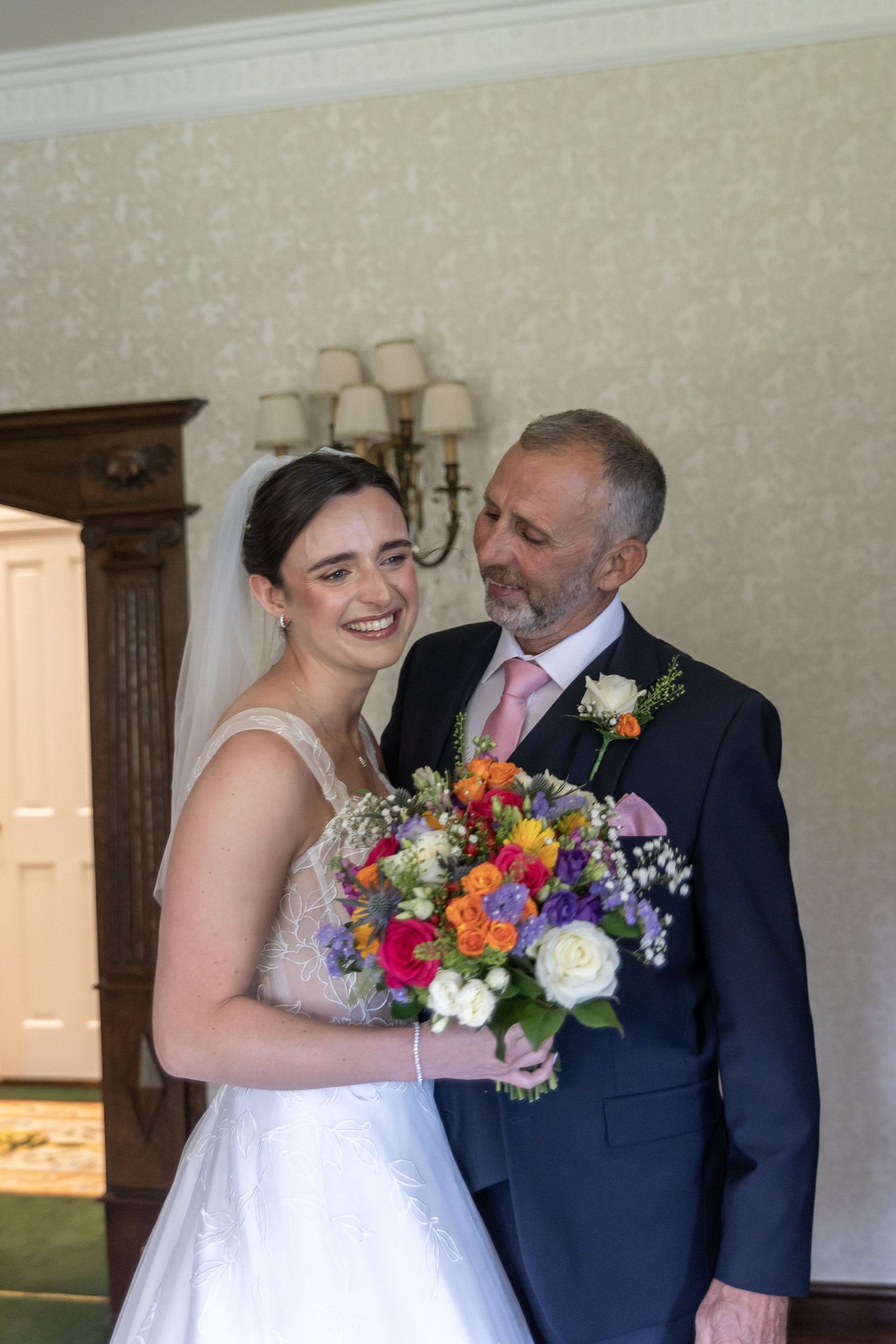 Bride in white gown holding colorful bouquet smiles with older man in navy suit and pink tie indoors.