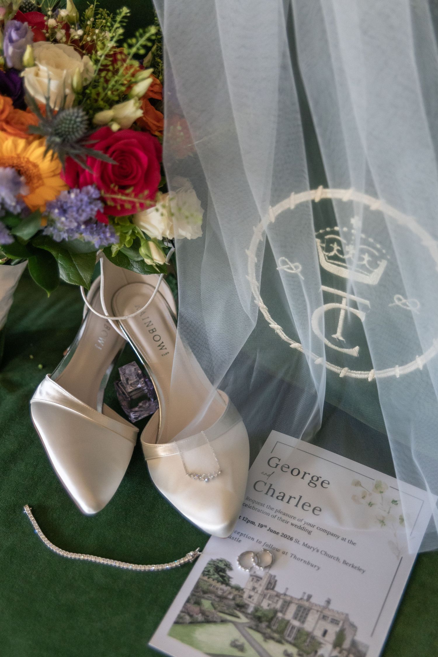 White bridal heels, wedding invitation for George and Charlee, diamond bracelet, and colorful bouquet on green surface.