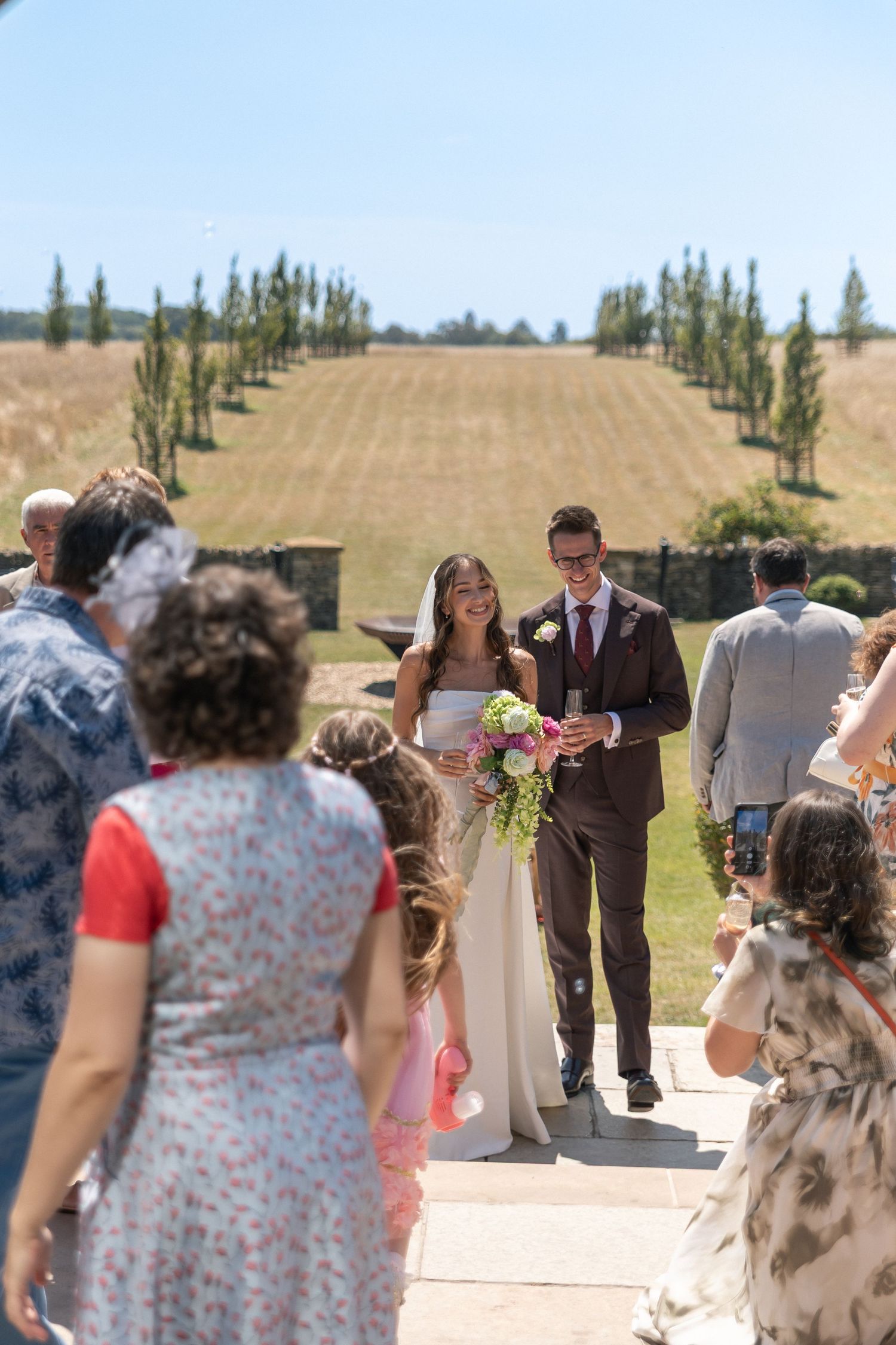 Bride and groom walk down the aisle outdoors at a sunny wedding ceremony surrounded by guests and cypress trees.