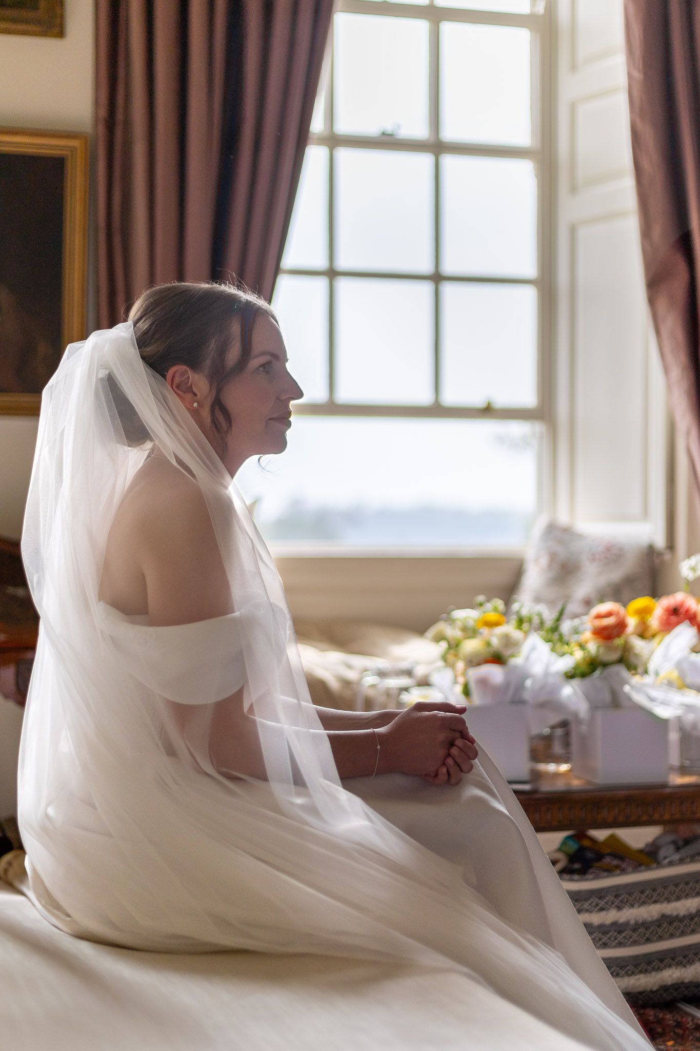 Bride in classic white gown sits gracefully by window with flowing veil and natural light.
