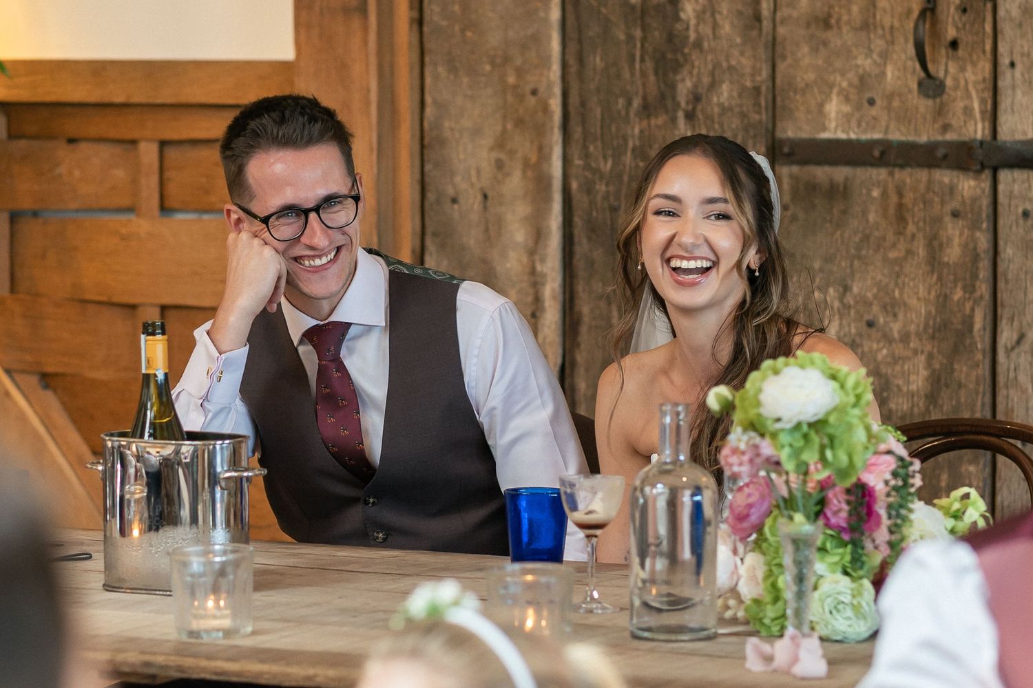 Bride and groom laughing joyfully at wedding reception table with floral centerpiece and champagne.