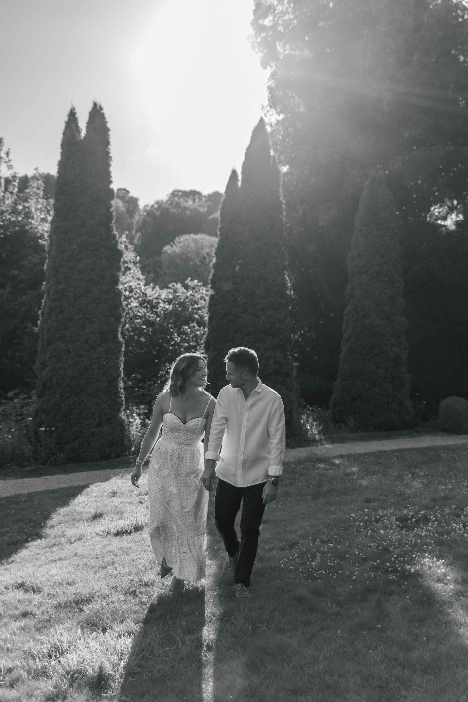 Black and white photo of a couple holding hands walking through a sunlit garden with tall cypress trees.