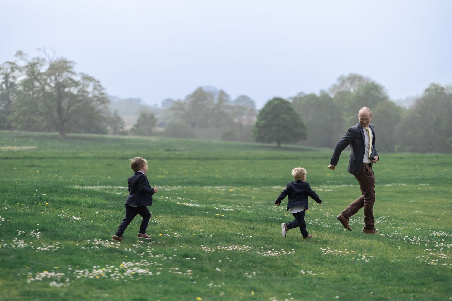 People running and playing together across a misty spring meadow filled with wildflowers on a cloudy day.