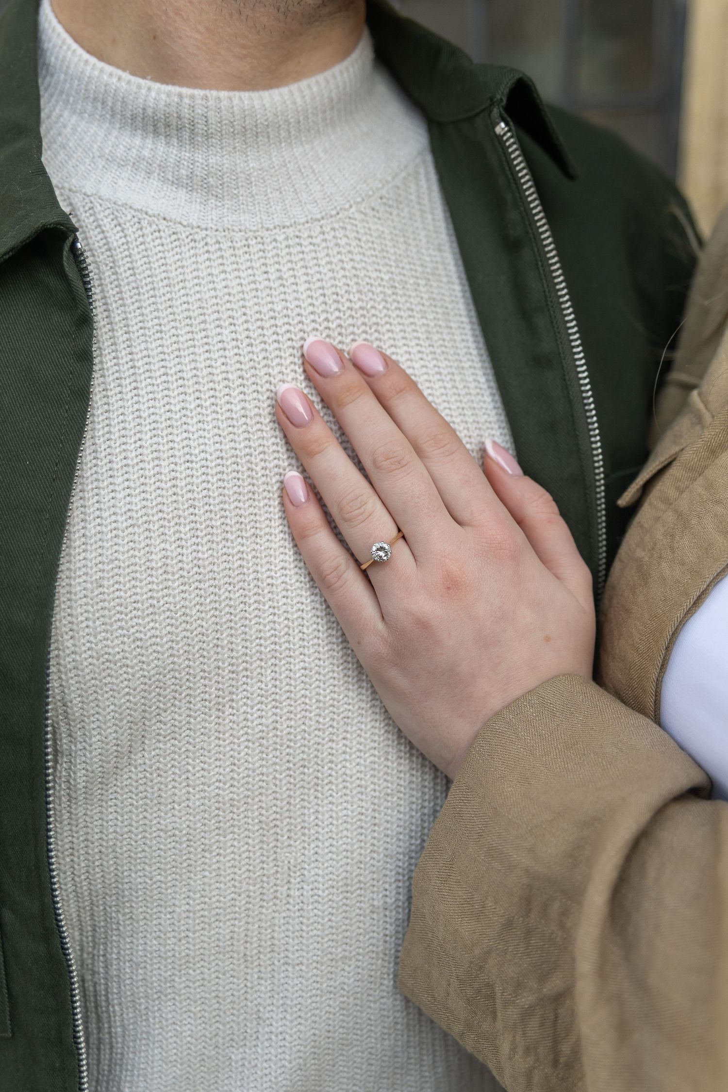 Close-up of a womans hand with a diamond engagement ring resting on a mans chest in a green jacket.