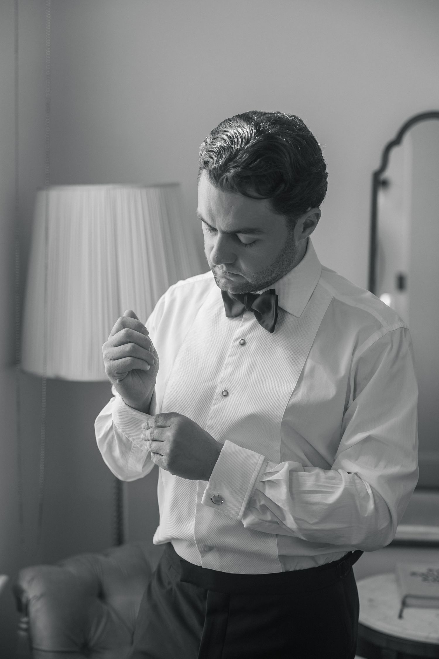 Black and white photo of a groom in a tuxedo shirt and bow tie fastening his cufflinks before his wedding at Fitzrovia Chapel