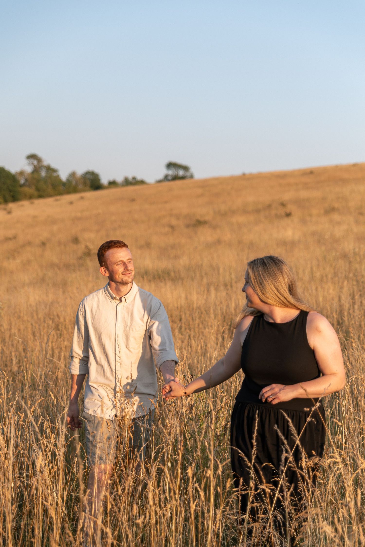 Couple holding hands in a golden wheat field at sunset, man in white shirt and woman in black dress.