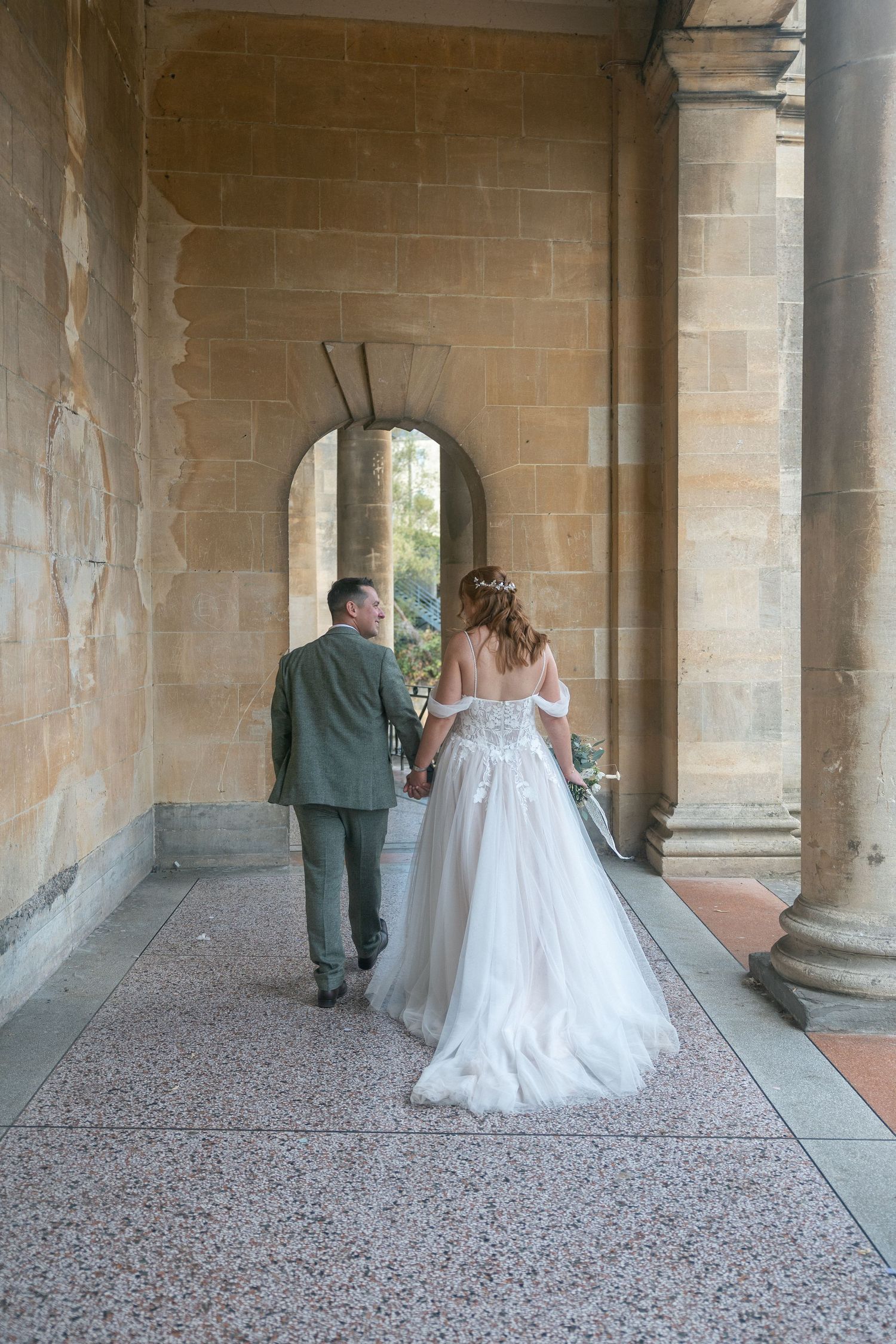 Bride and groom walking hand in hand through elegant stone archway colonnade in wedding attire in Parade Gardens, Bath