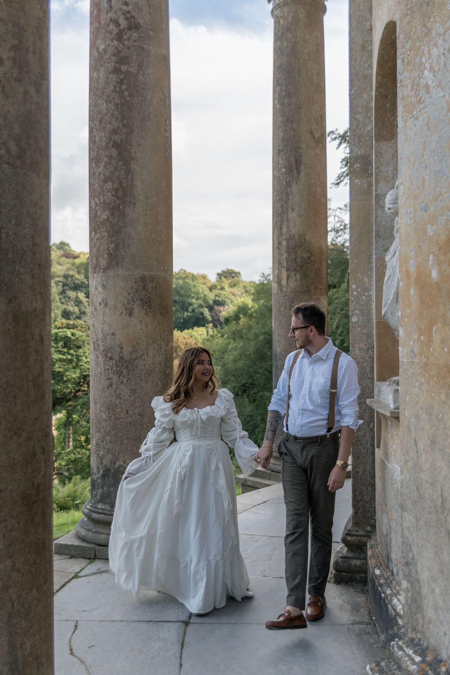 Bride and groom walk hand in hand between stone columns at a historic venue, surrounded by lush greenery.