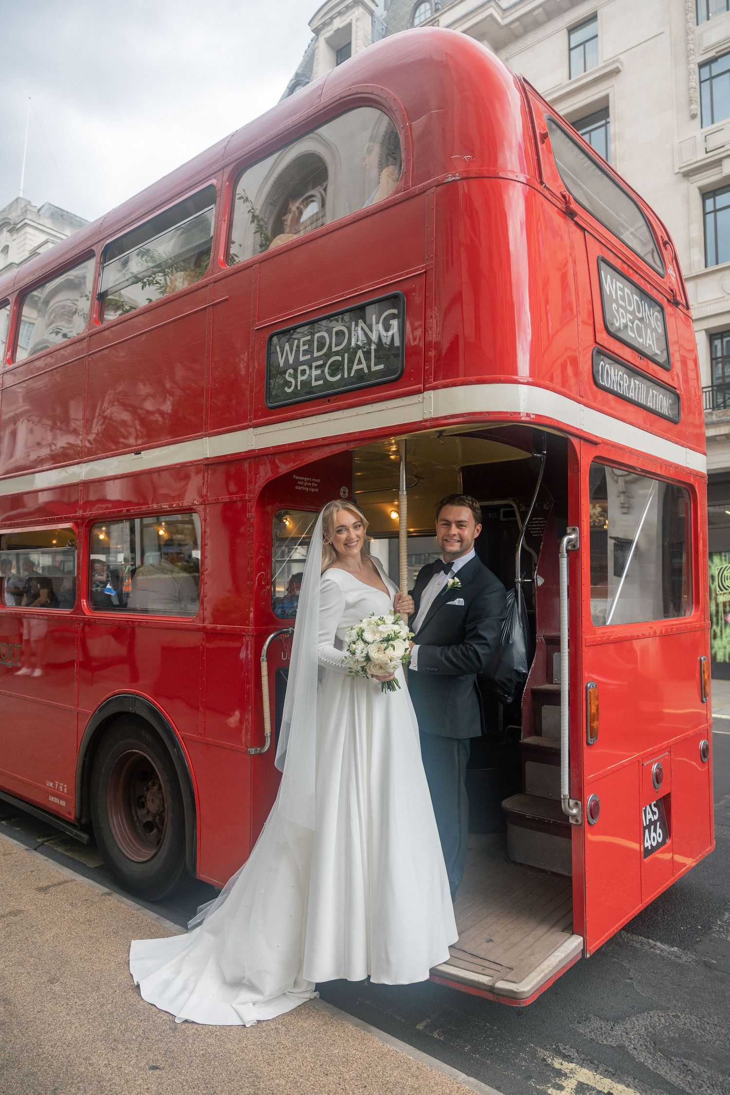 Bride and groom posing by a red London double-decker Wedding Special bus, bride holding white bouquet.