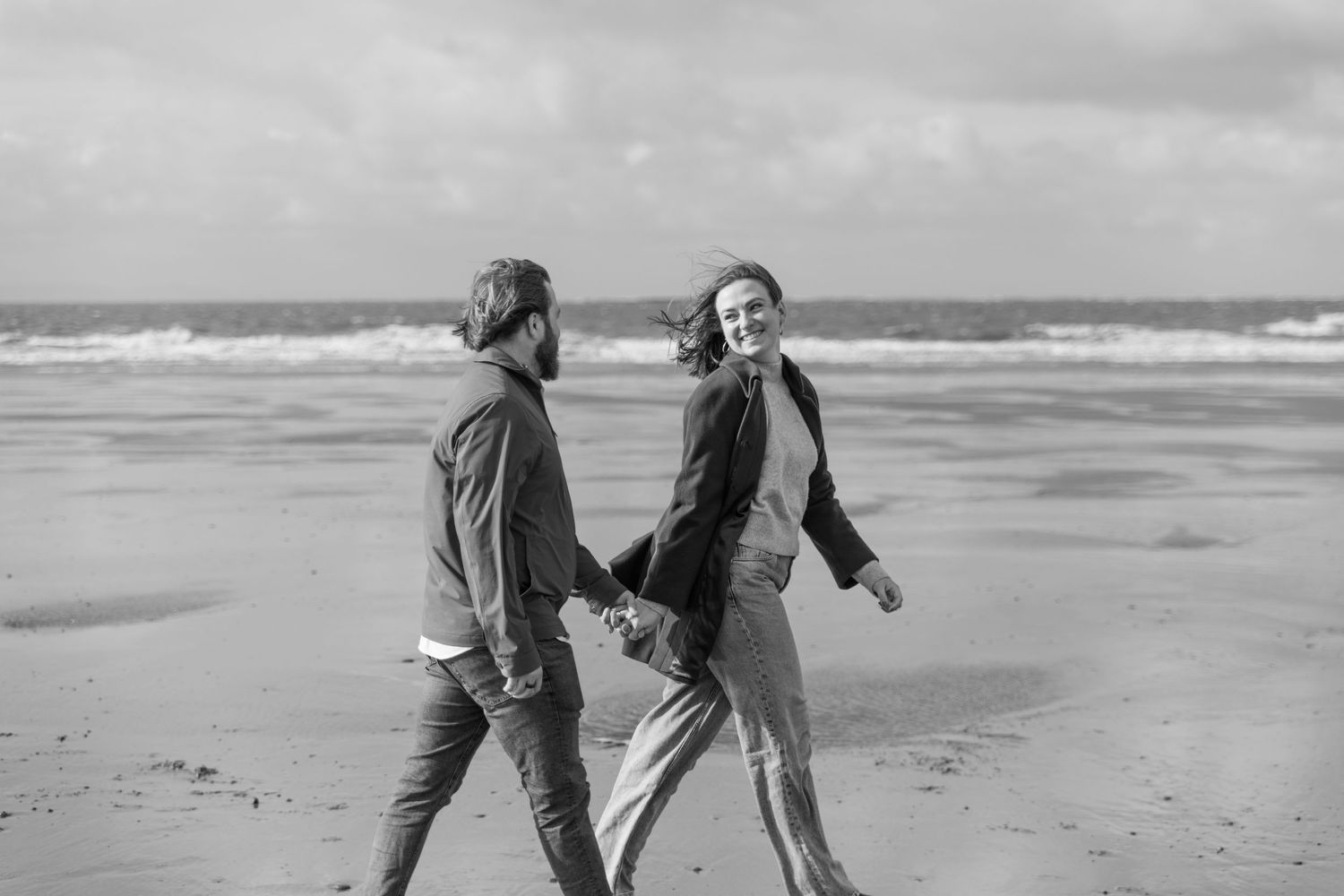 Two people stroll along a misty beach shore during an overcast day.