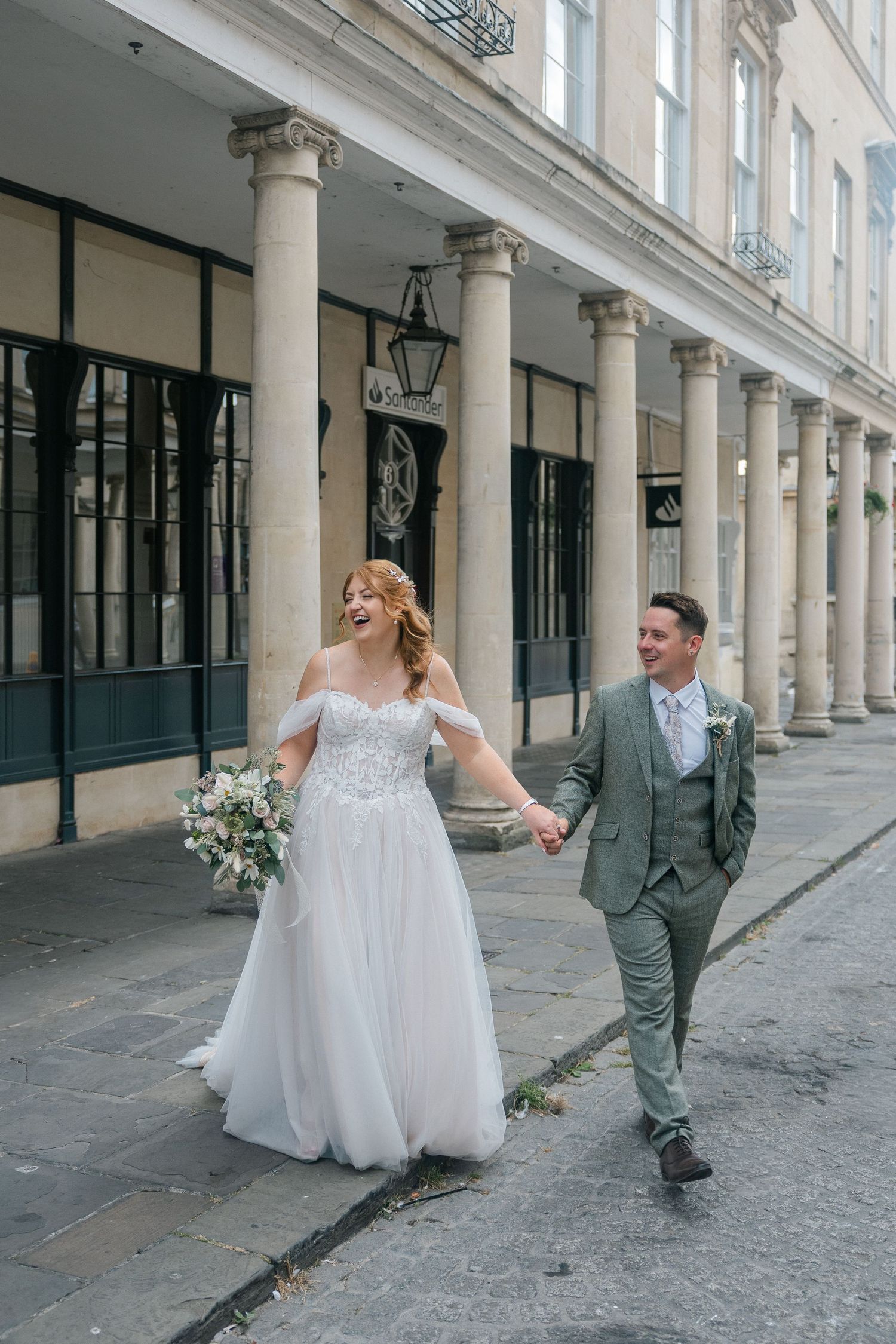 A joyful bride and groom walk hand-in-hand past elegant stone columns on their wedding day.