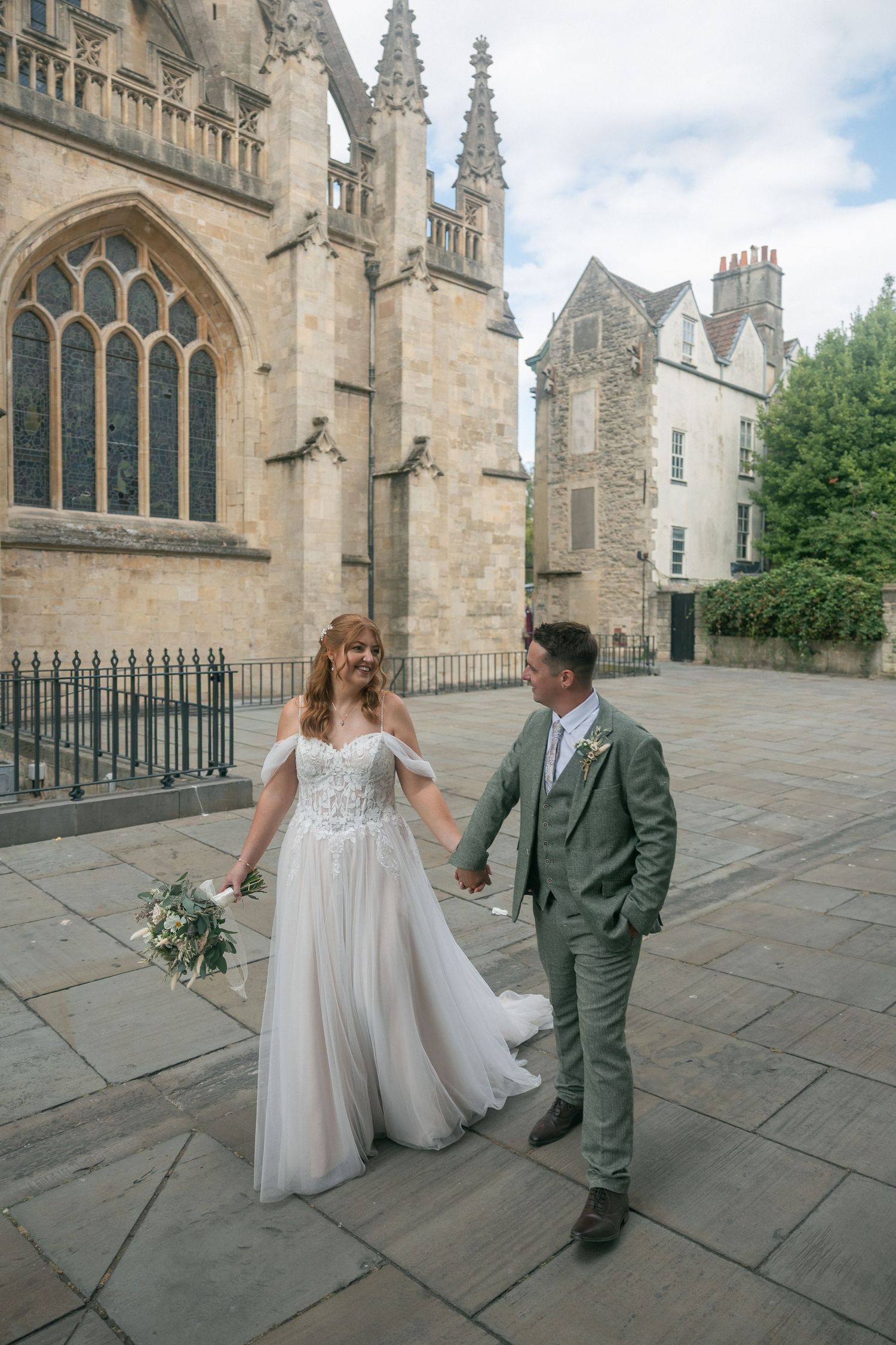 Bride and groom holding hands walking outside Bath Abbey, bride in white gown, groom in sage green suit.