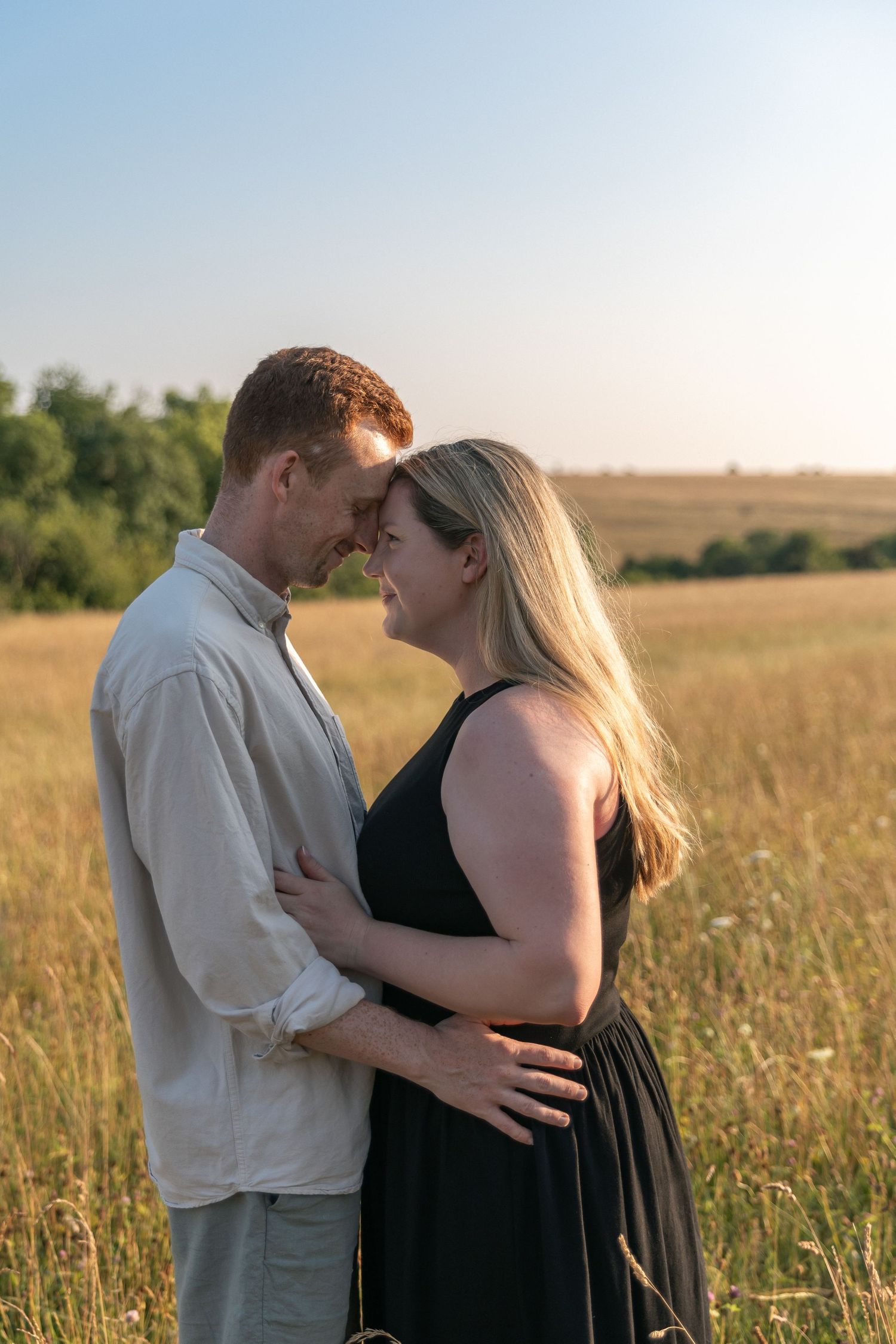 A couple shares a romantic moment in a golden field at sunset, touching foreheads and embracing tenderly.