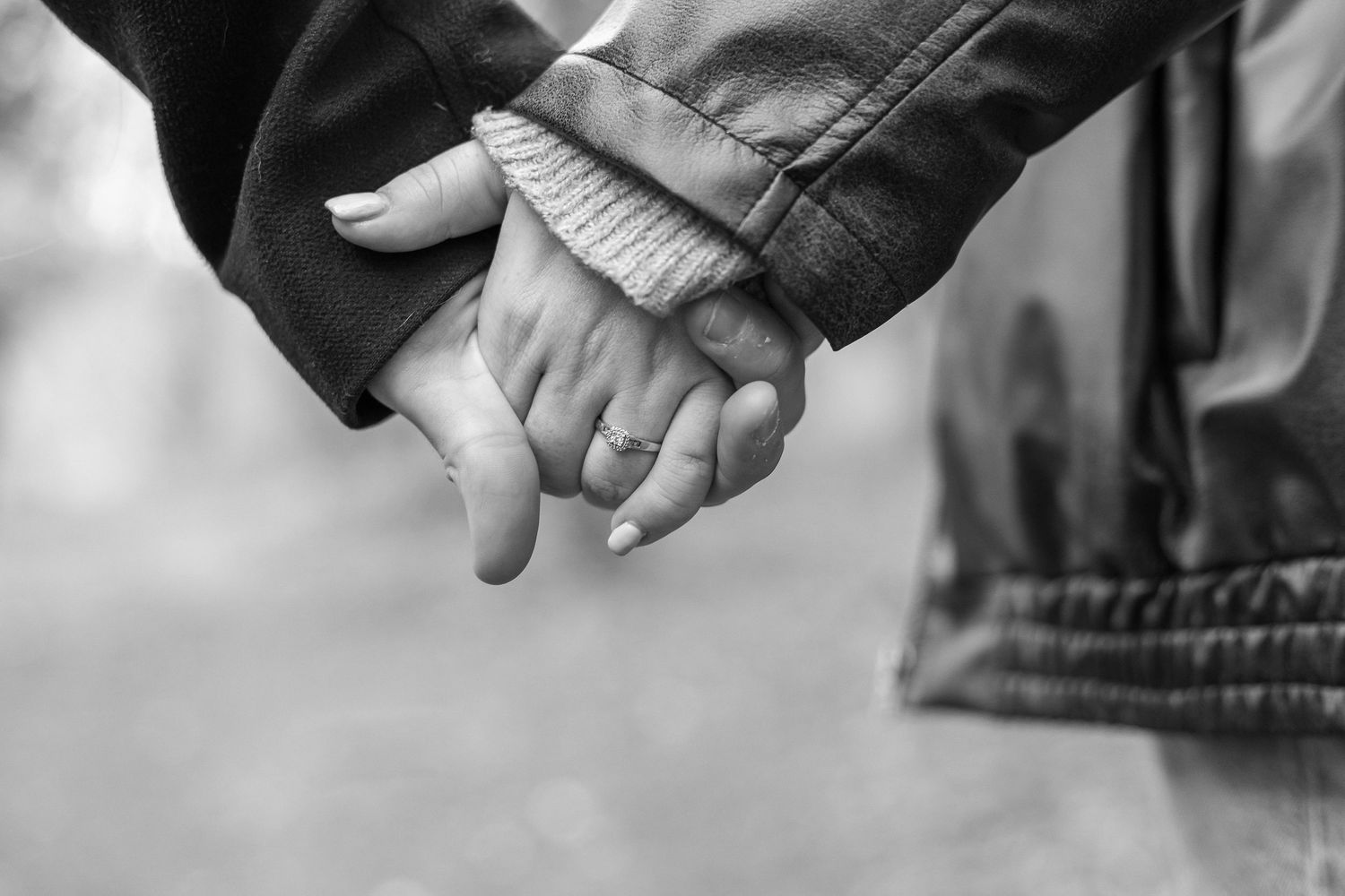 Black and white photo of a couple holding hands, showcasing a ring and painted nails in a romantic outdoor setting.
