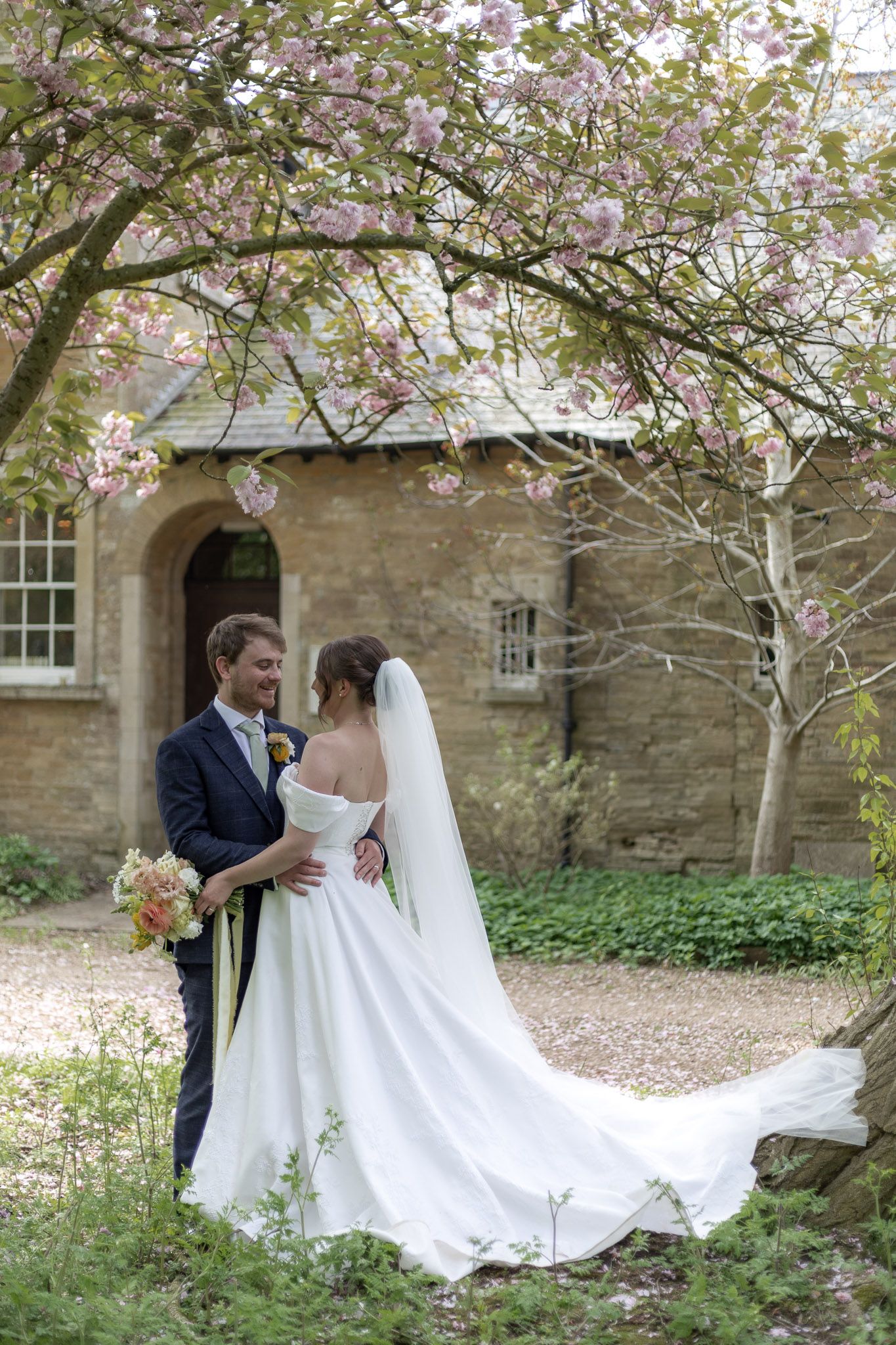 A flowing white wedding dress trails behind the couple as they pose beneath blooming trees.