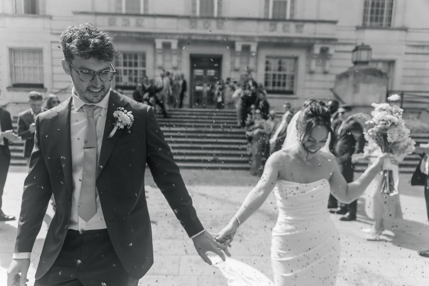Black and white photo of a newlywed couple holding hands and smiling as they walk away from a grand building.