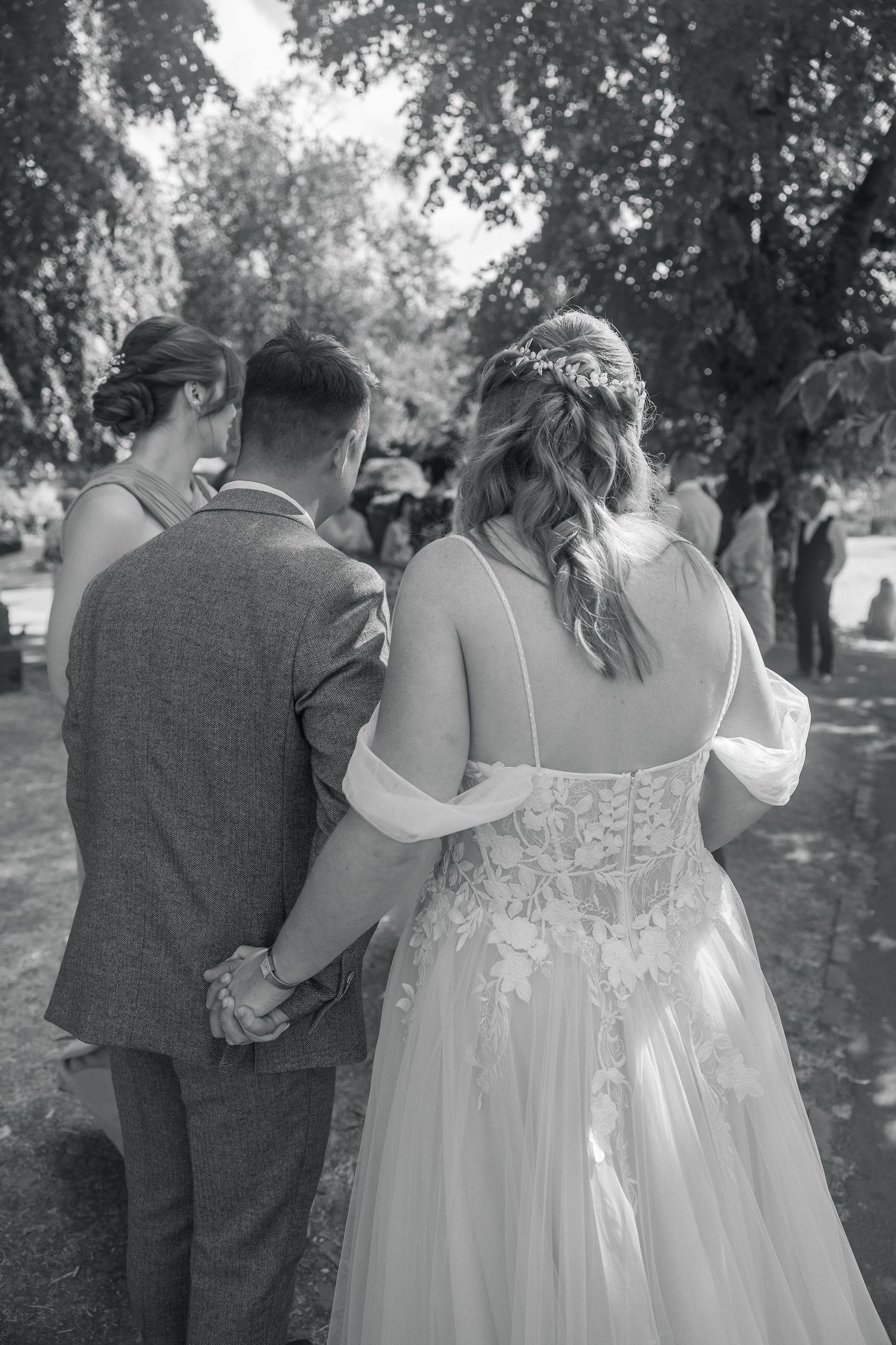 Black and white photo of a bride and groom holding hands, walking outdoors, bride wearing lace wedding gown with floral hair piece.