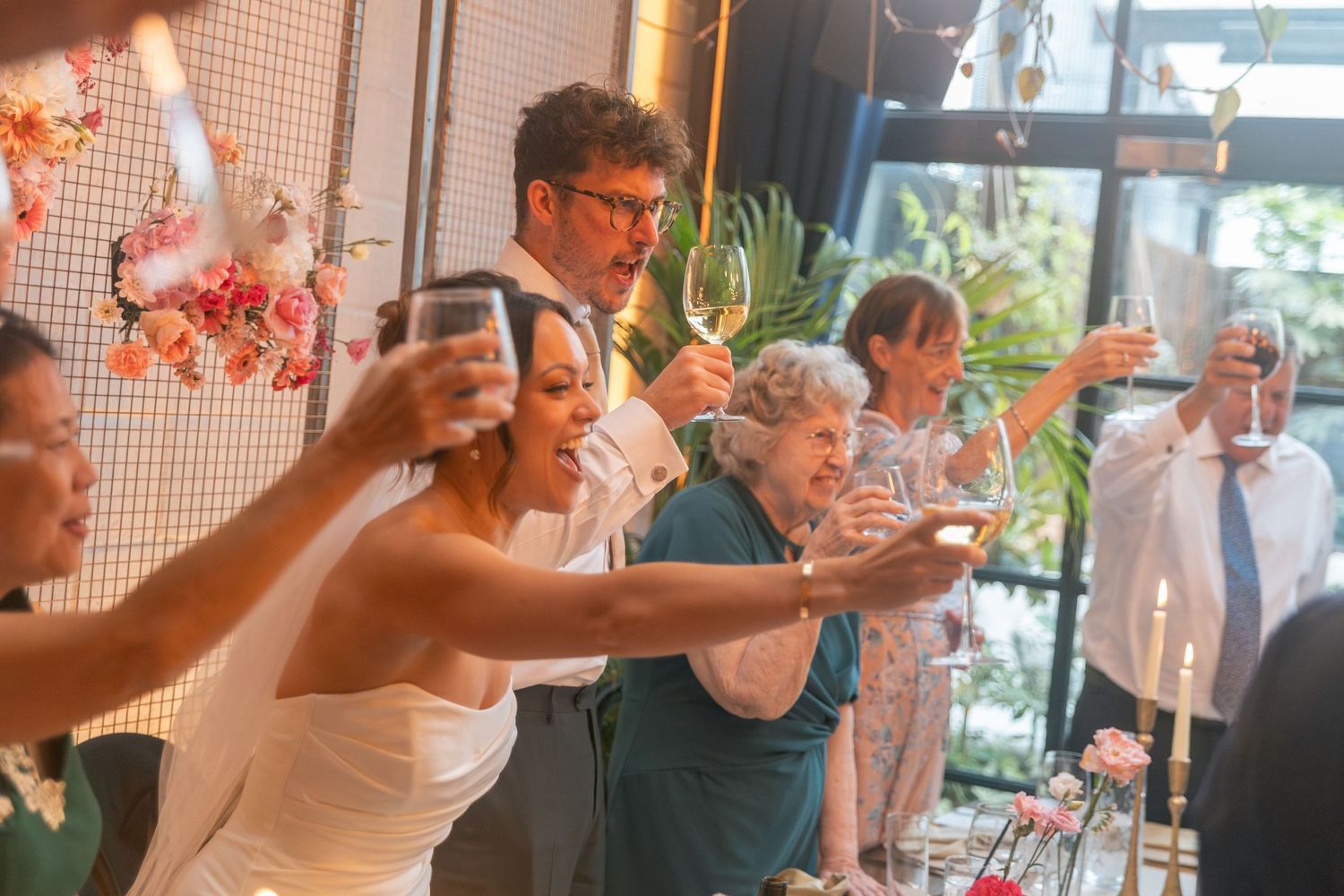 Wedding guests raising glasses in a joyful toast, with bride in white dress celebrating at reception at The Hackney.