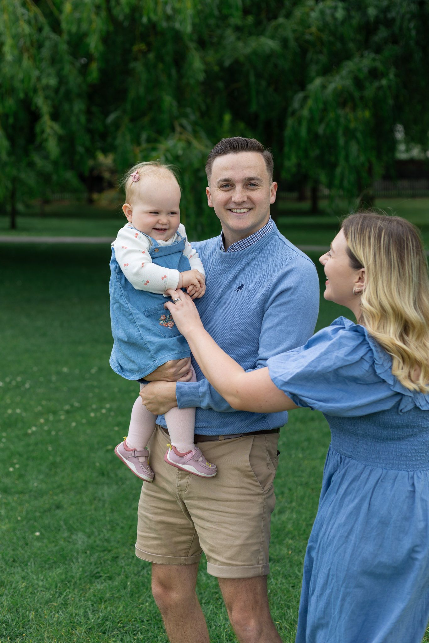 Family wearing coordinating blue and khaki outfits poses together on green lawn with willow trees in background.