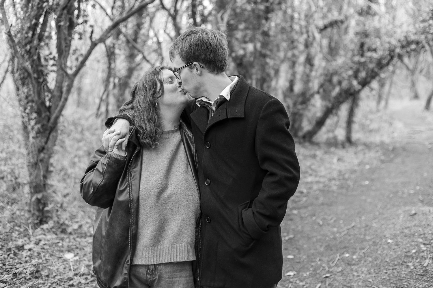 Black and white photo of a couple sharing a kiss on a forest path surrounded by bare autumn trees.