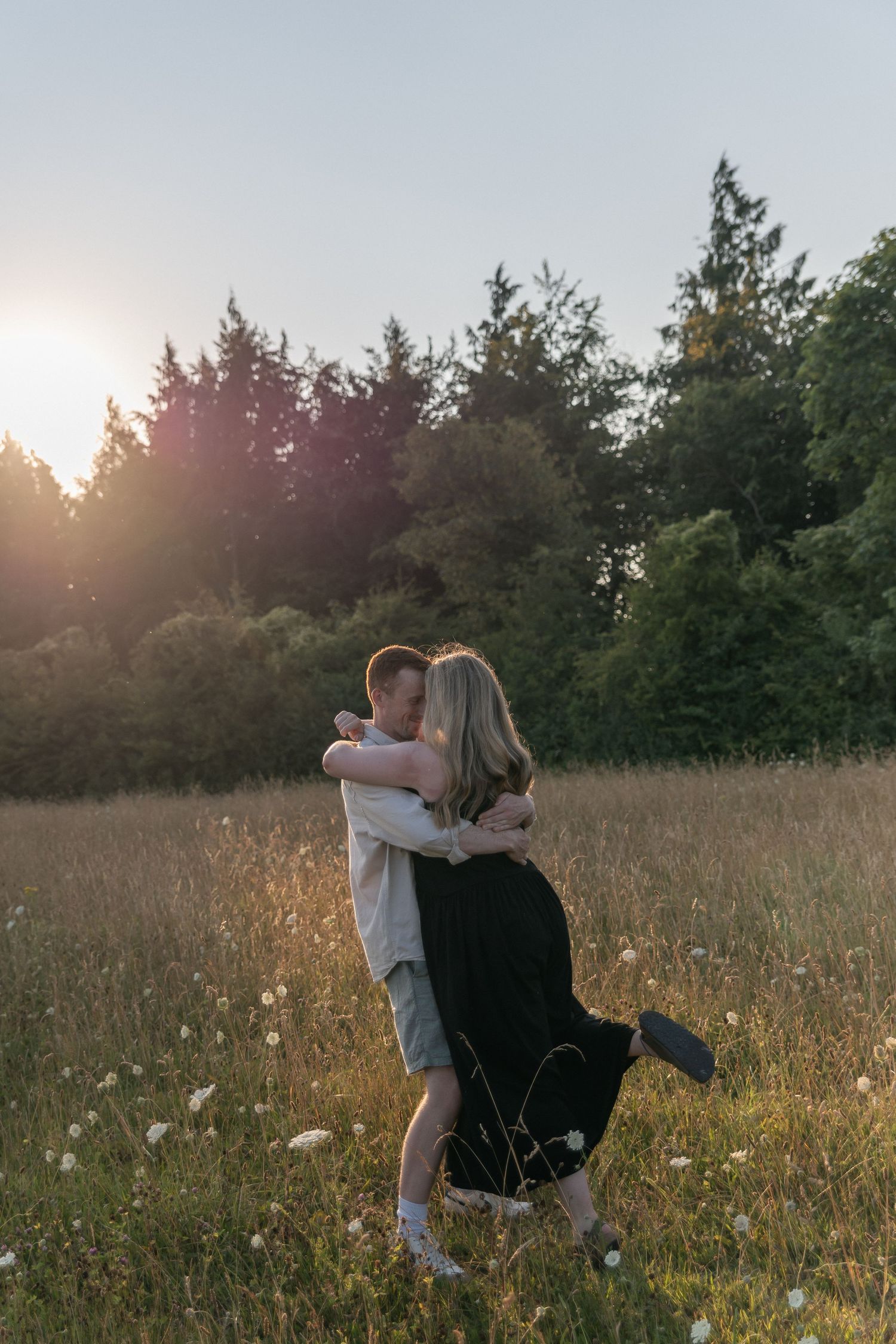 Couple embracing in a sunlit meadow with wildflowers and trees at golden hour sunset.