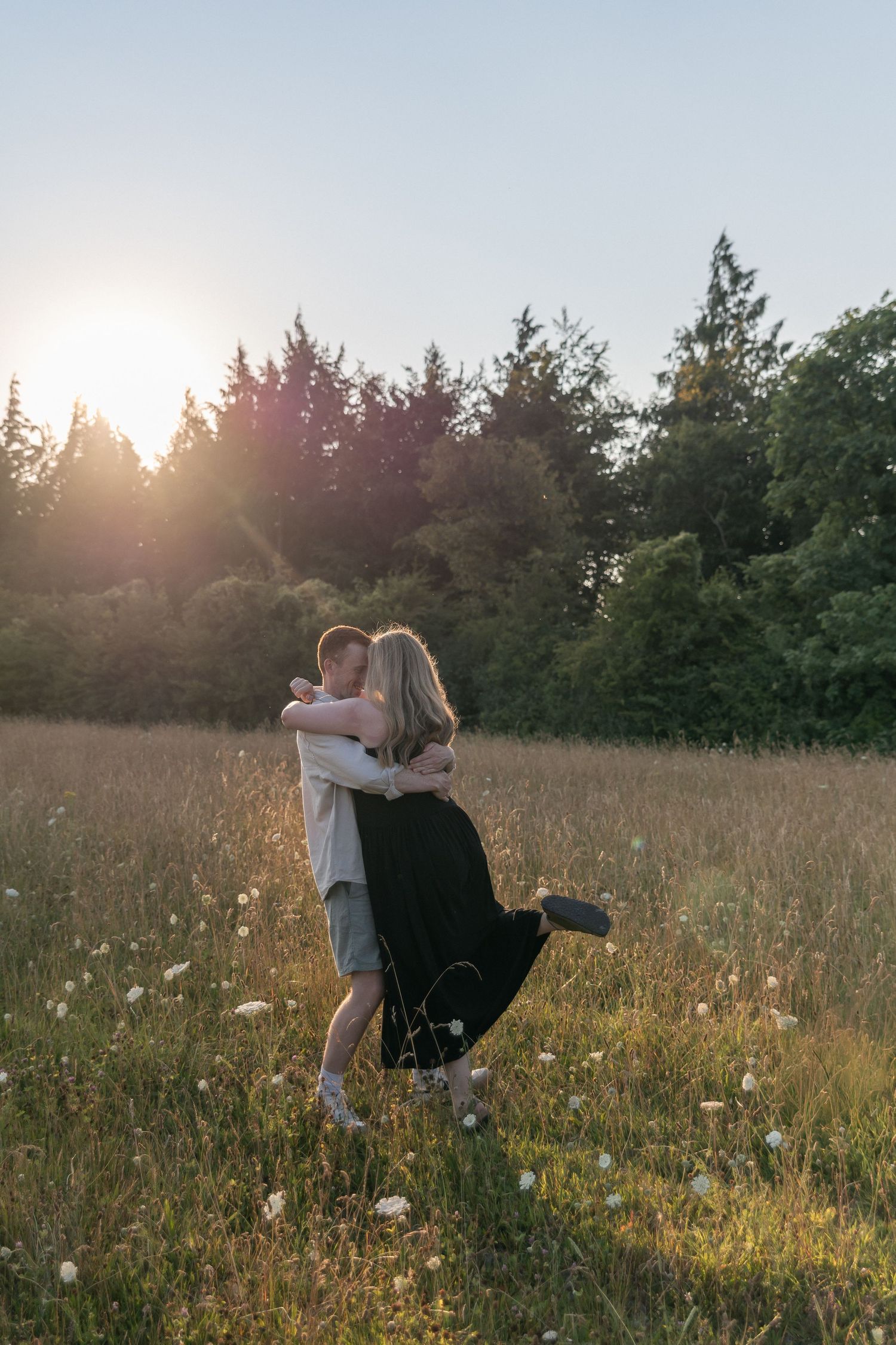 Couple embracing in a sunlit meadow with wildflowers and trees at golden hour.