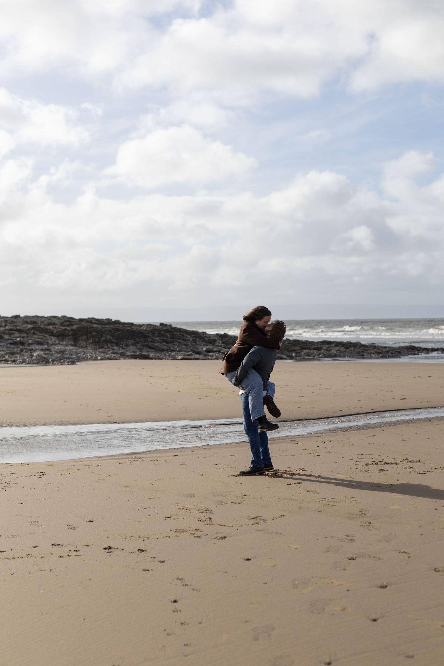 Two people playfully interact on a wide sandy beach with waves and rocky cliffs in the distance.