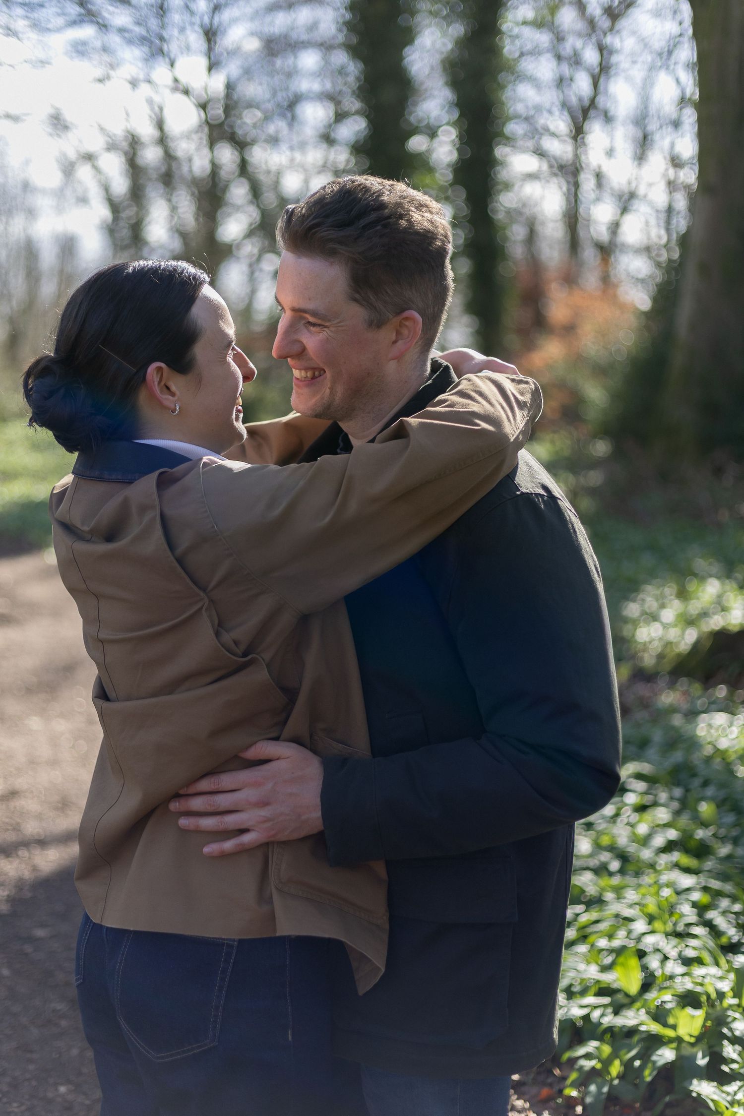 A couple embraces warmly outdoors in a wooded area with natural lighting and greenery in the background.