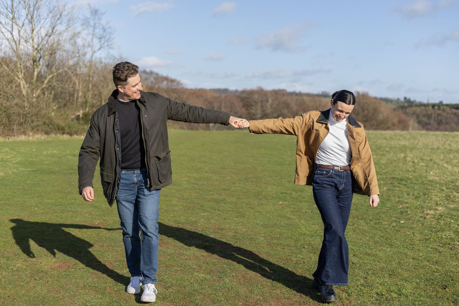 A person wearing blue jeans and a jacket walks through a grassy field on a sunny day.