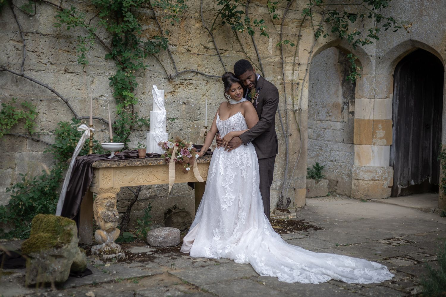 Bride and groom pose together in lace wedding dress and dark suit at rustic stone  outdoor ceremony at Sudeley Castle