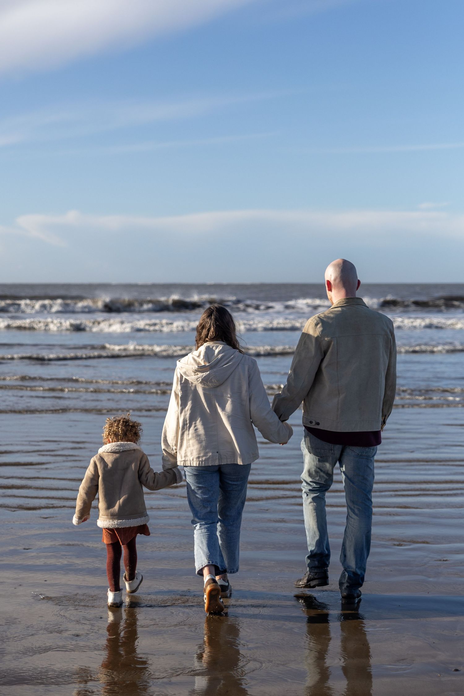 A group walks together along a wet sandy beach at low tide with reflections visible in the shallow water.