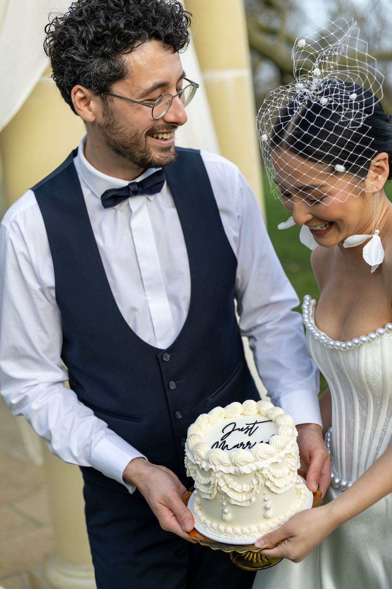 A newlywed couple shares joyful moments while holding their white wedding cake decorated with delicate frosting details.