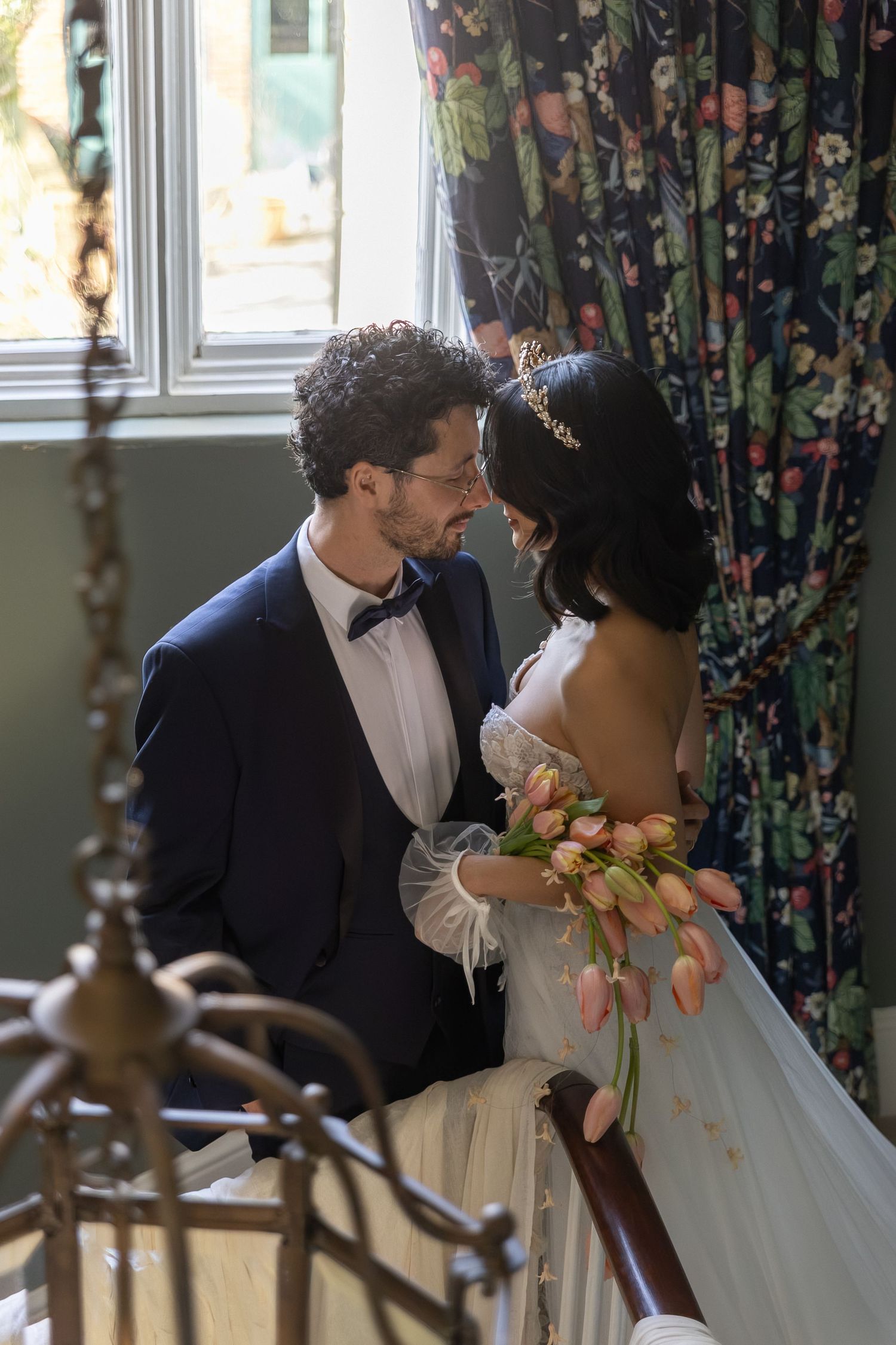 A couple in formal wedding attire share an intimate moment near a window with floral curtains.